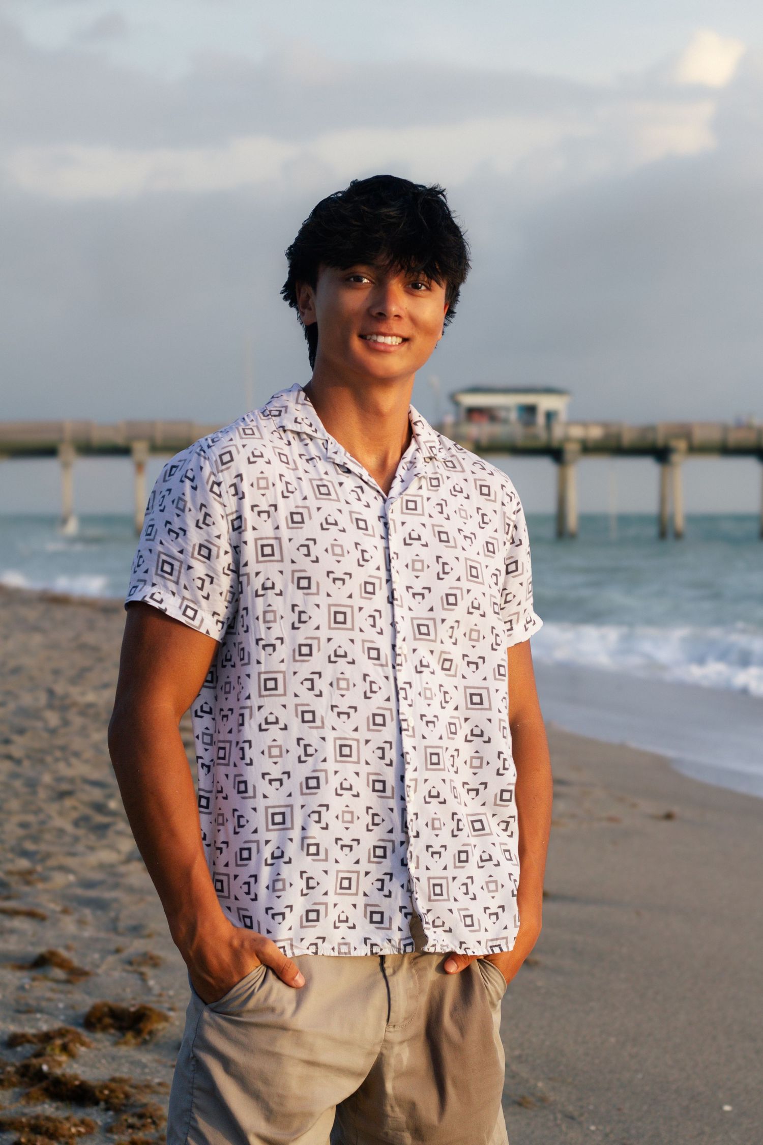 Young man smiling on beach wearing patterned shirt, hands in pockets, pier and ocean waves in background.