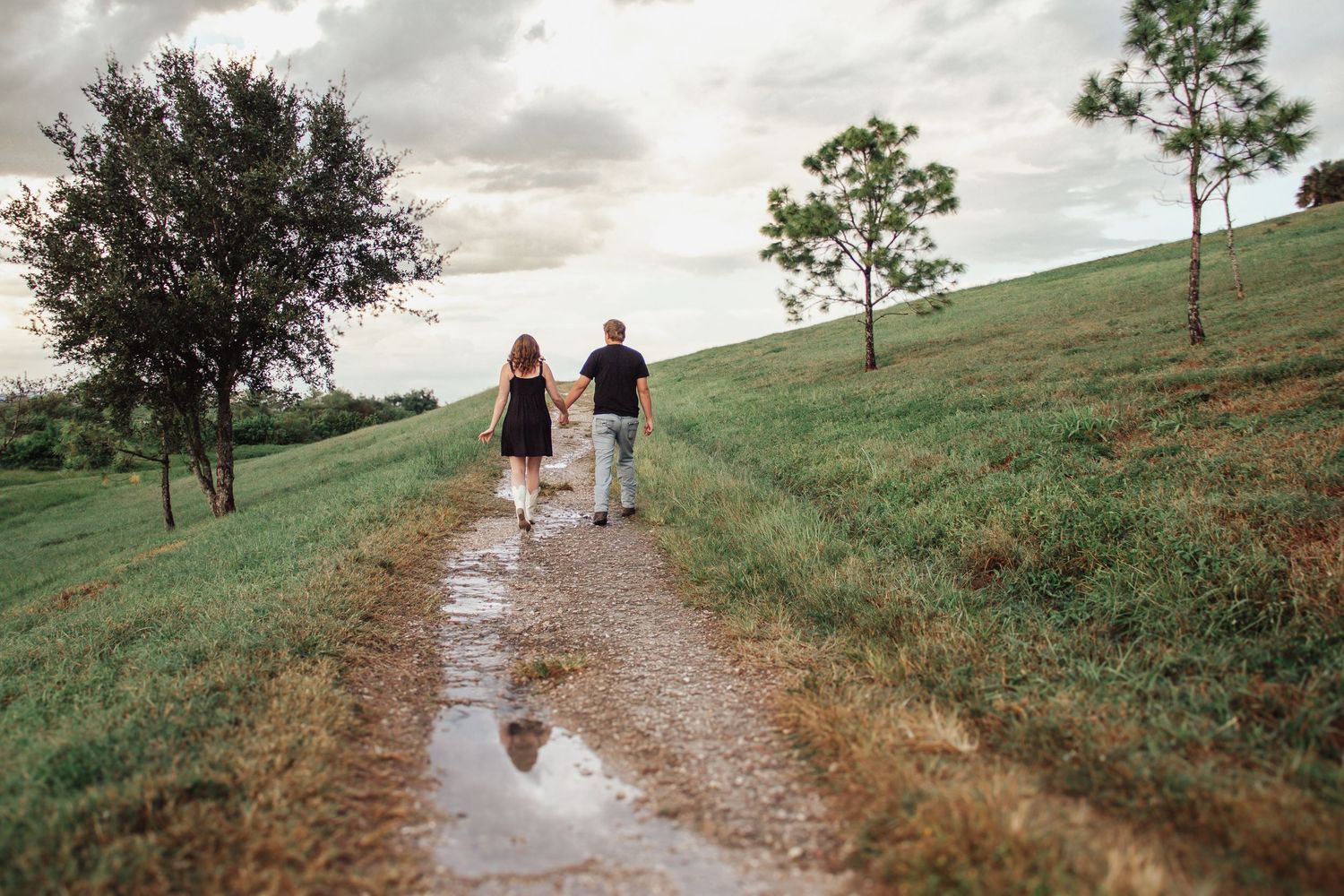 Couple walking hand-in-hand on a gravel path through lush green hills after rain, with puddles reflecting the cloudy sky.