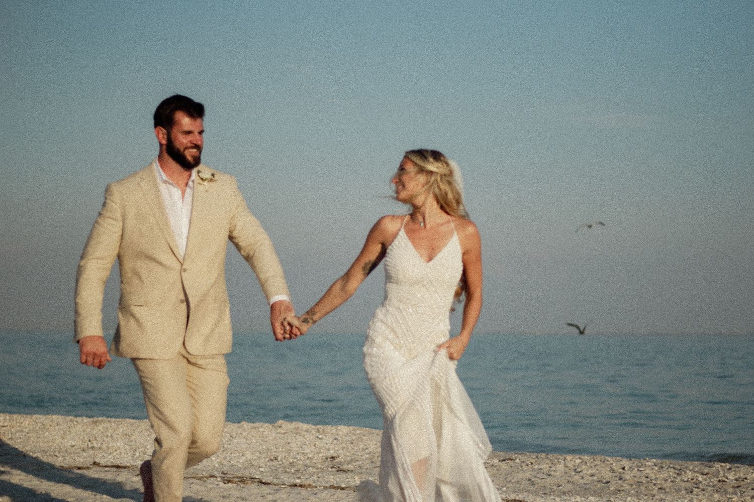 Bride and groom holding hands walking on a sandy beach in wedding attire with ocean backdrop.