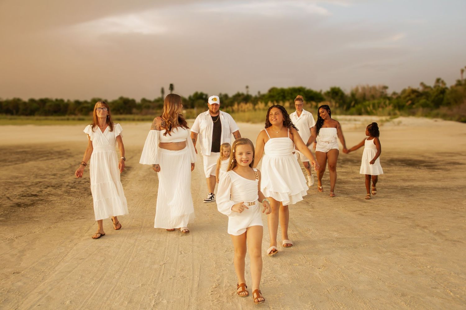 Family in white outfits walking on sandy beach at golden hour, children running ahead joyfully.
