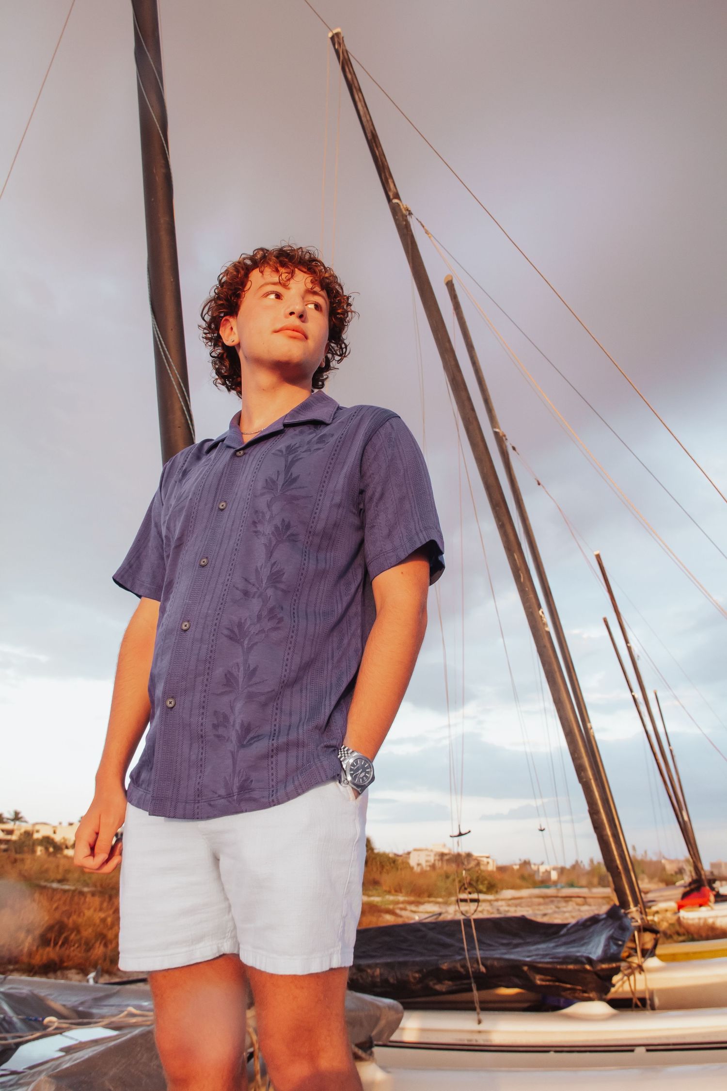 Young man with curly hair in blue shirt and white shorts standing by sailboat masts at sunset.