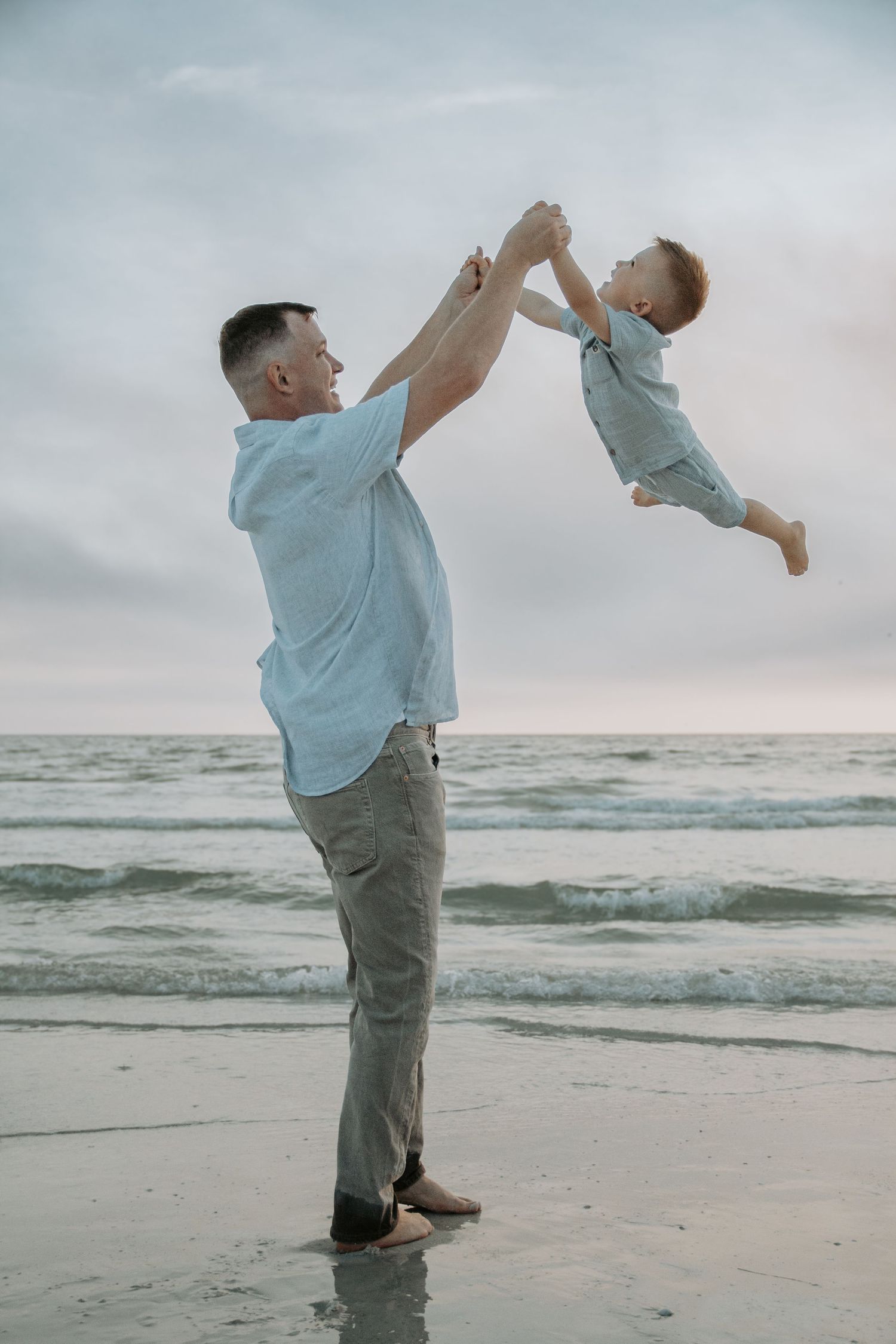 Father joyfully lifting toddler high in the air on a serene beach at dusk, both dressed in light blue.