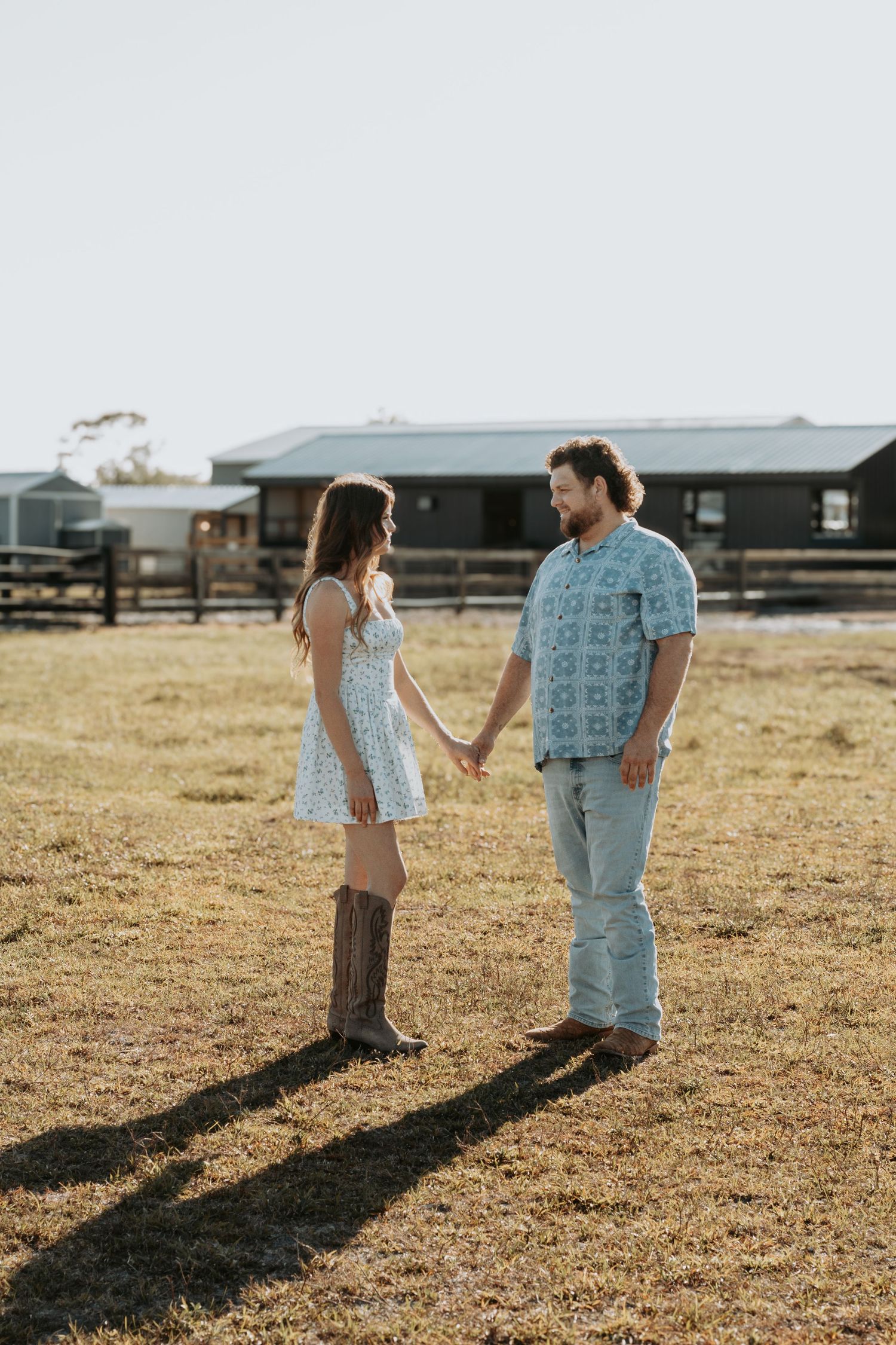 A couple holds hands in a sunny farm field, woman in white dress and cowboy boots, man in blue patterned shirt.