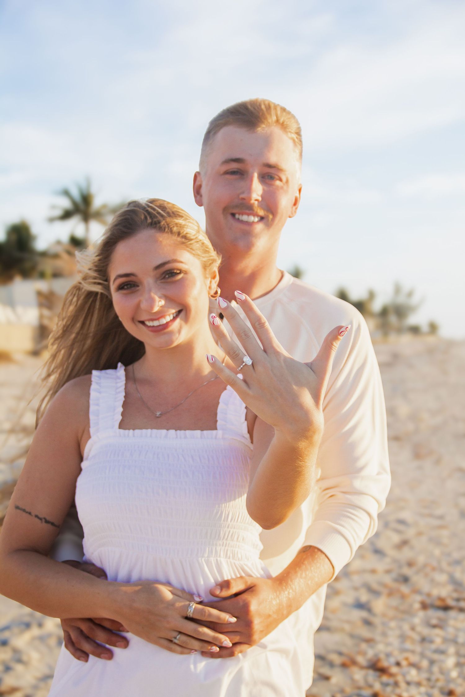 Happy couple on beach showing engagement ring, woman in white dress, man in cream shirt, palm trees background.