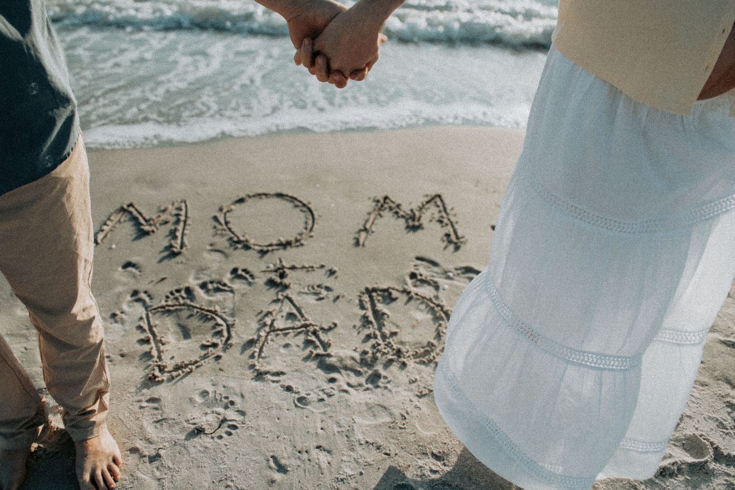 Couple holding hands on beach with MOM and DAD written in sand near ocean waves.