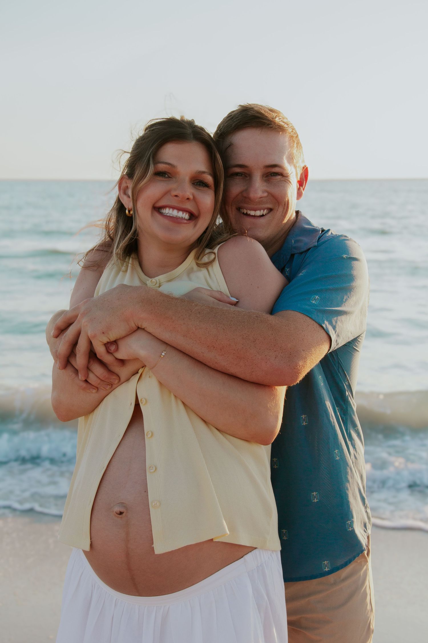 Happy couple embracing on beach at sunset, showing pregnant belly, smiling at camera.