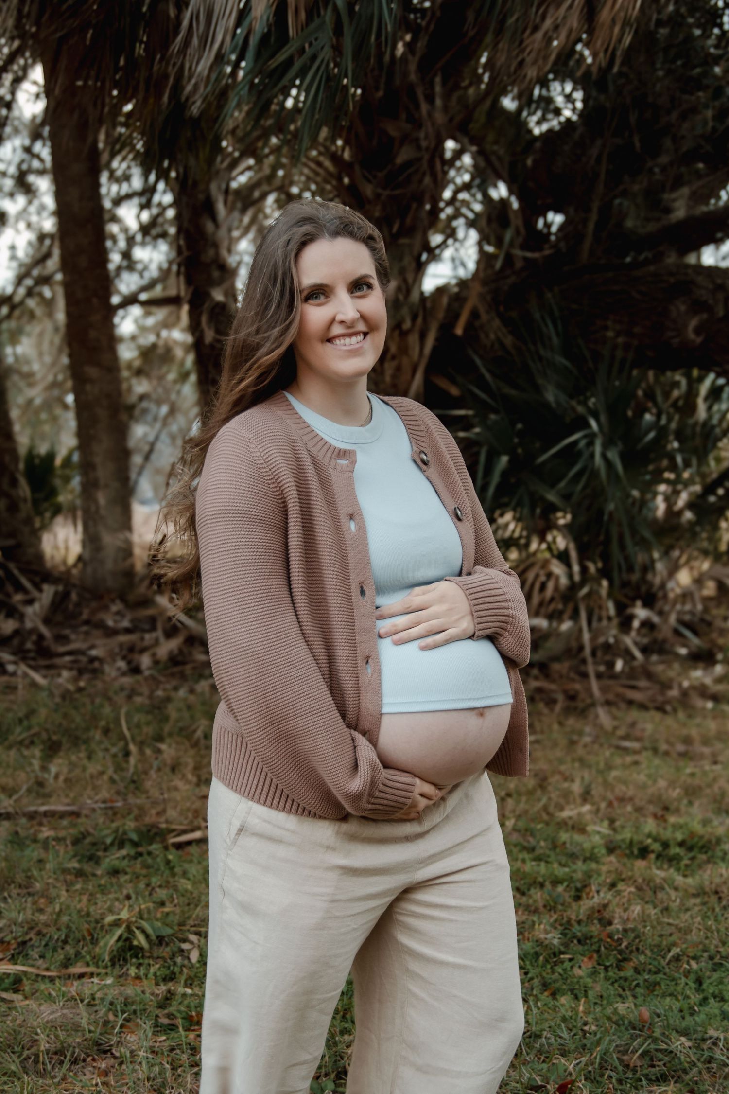 Smiling pregnant woman in brown cardigan and light blue top cradling baby bump outdoors among lush green trees.