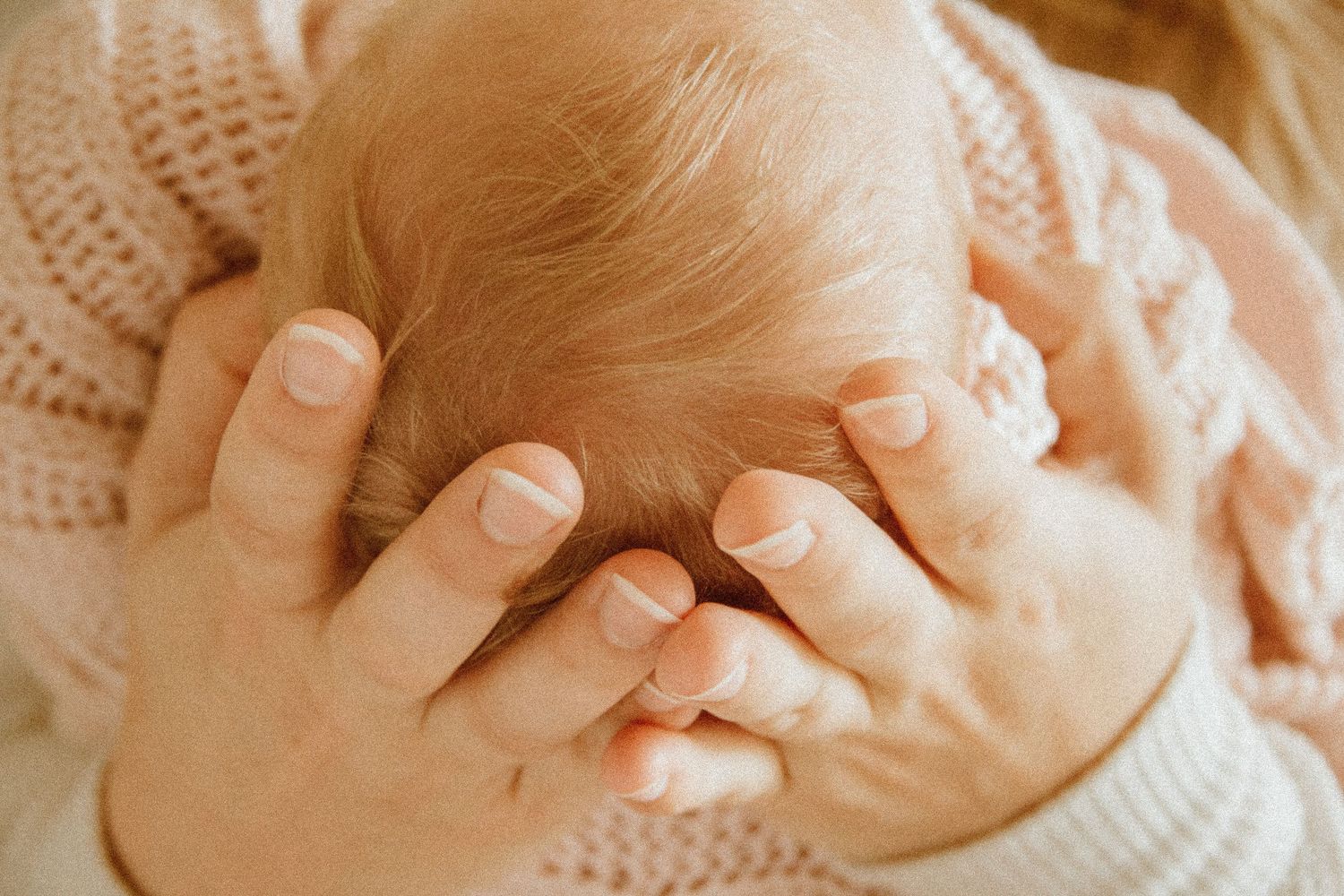 Close-up of a newborn baby's head cradled gently in a parent's hands, wearing a soft pink knit outfit.