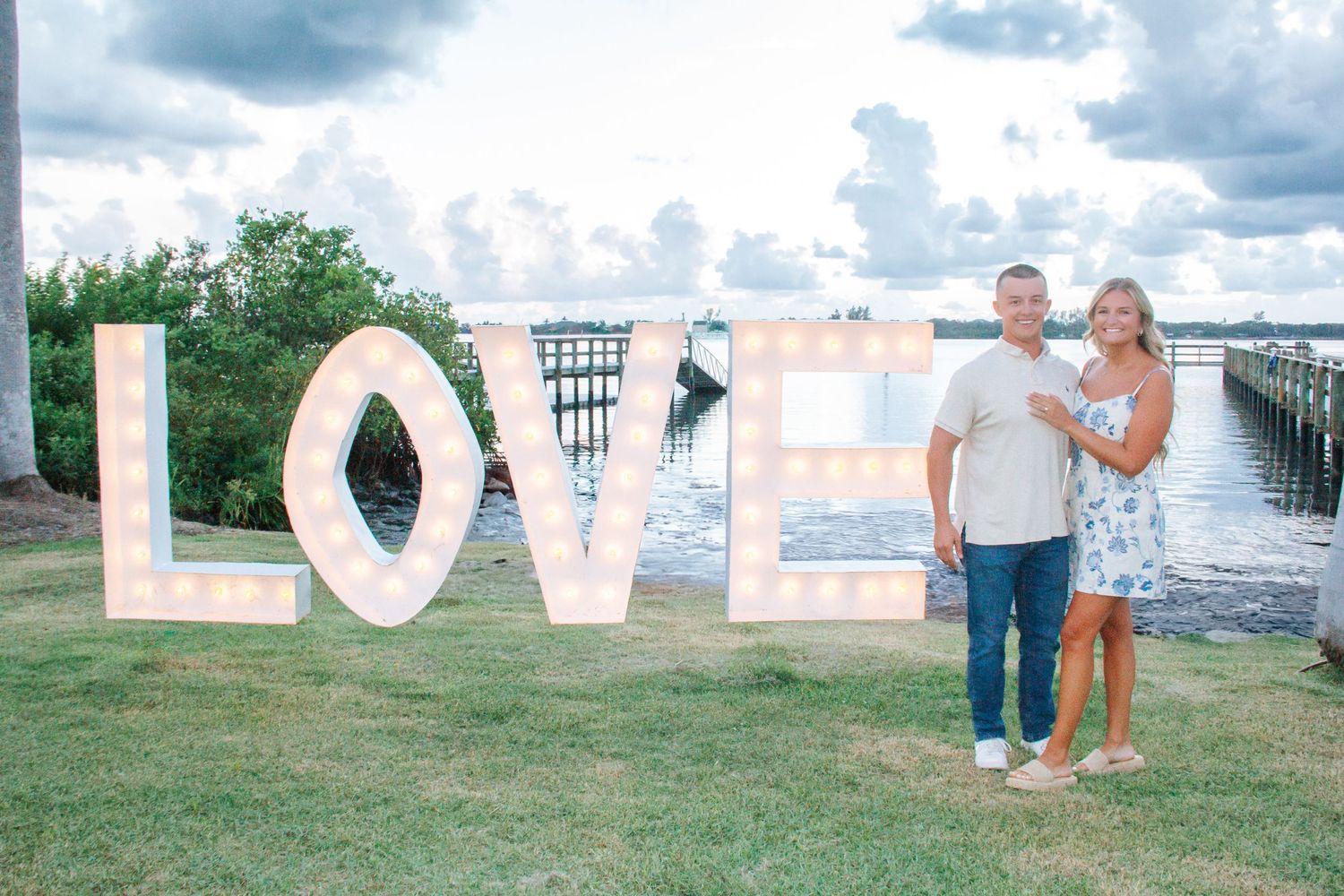 Couple posing in front of large illuminated LOVE letters on waterfront lawn with cloudy sky backdrop.
