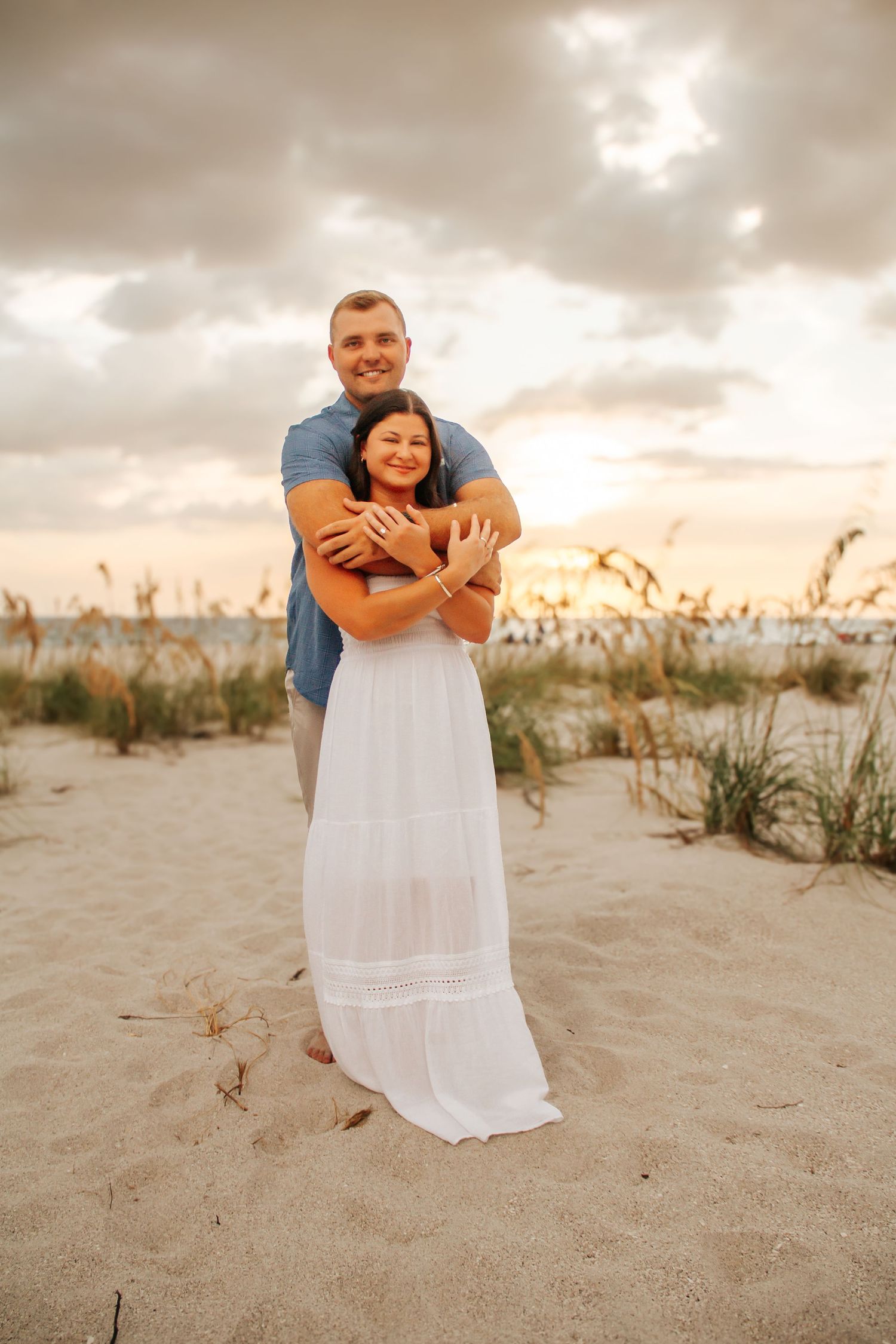 Couple embracing on sandy beach at sunset, man in blue shirt hugging woman in white maxi dress.