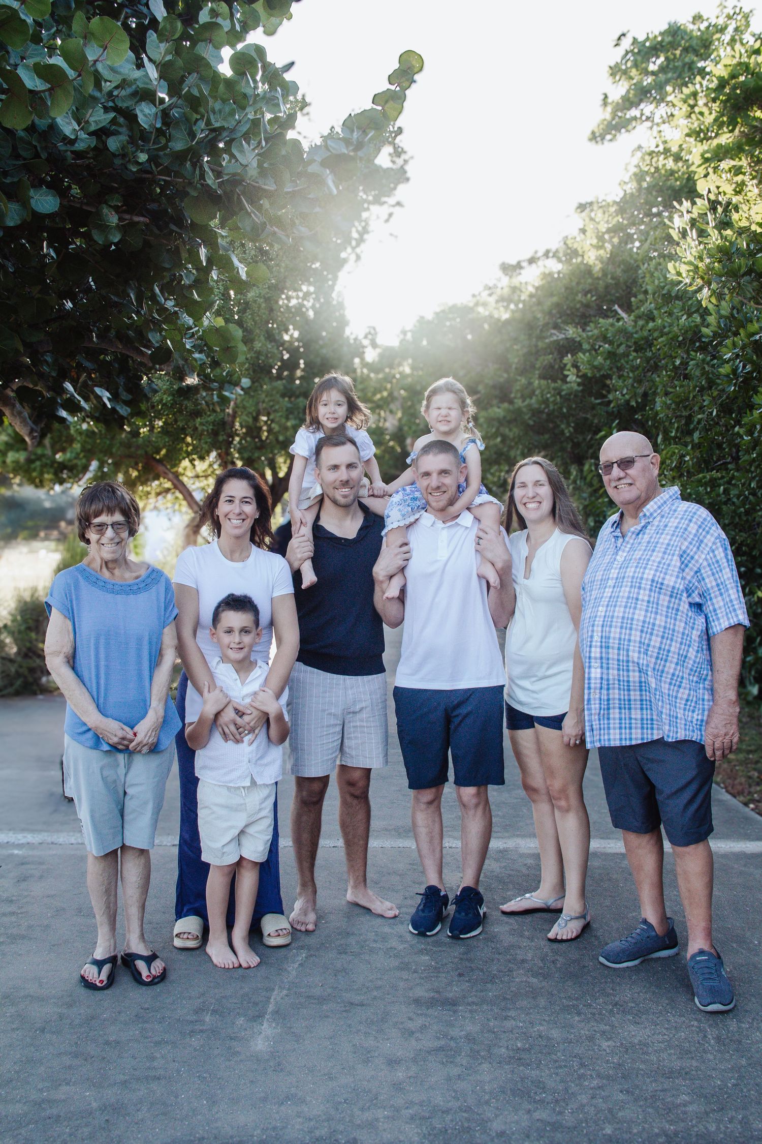 Multi-generational family portrait outdoors with trees and sunlight, featuring grandparents, parents, and young children.