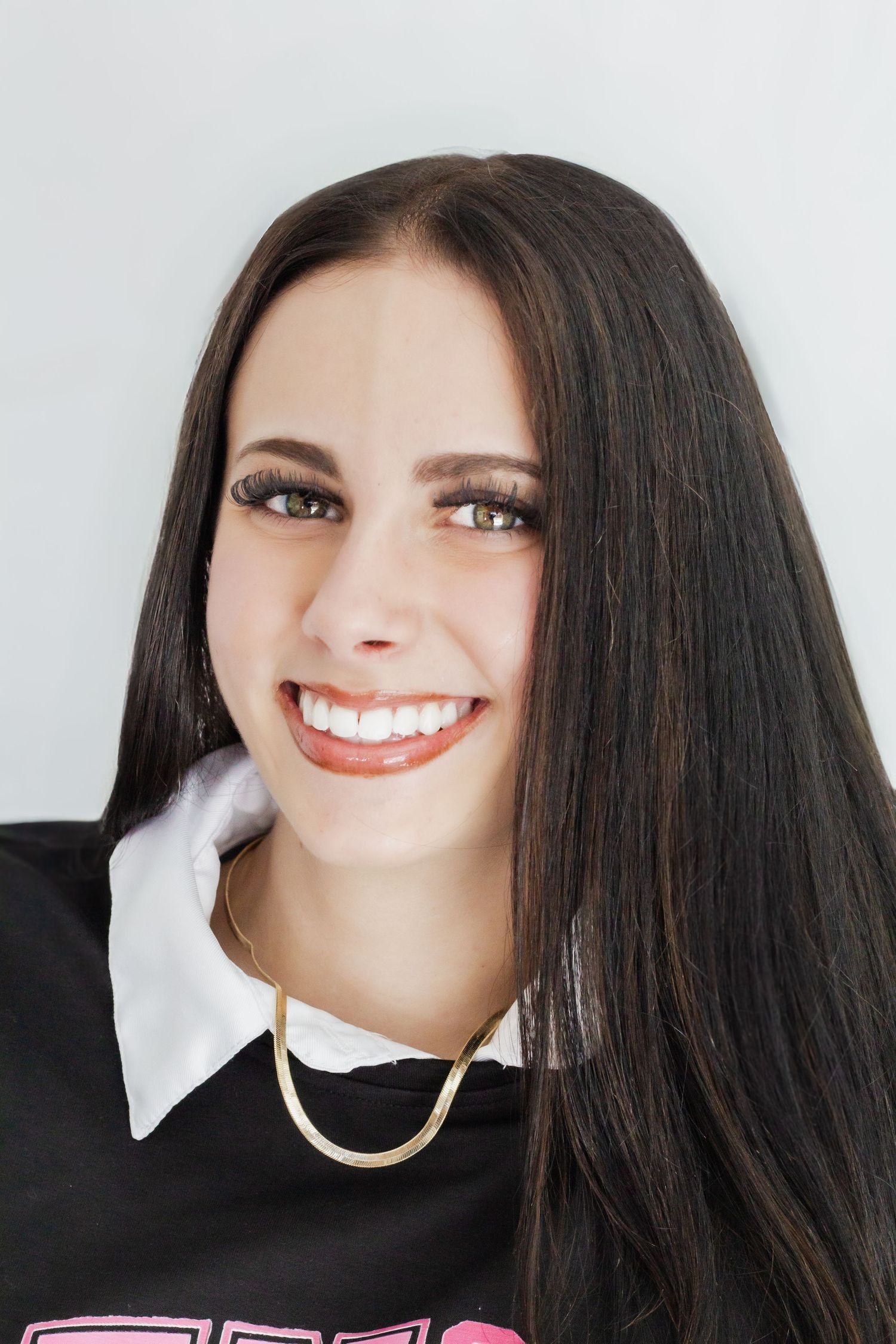 Smiling young woman with long dark hair wearing a black top with white collar and gold chain necklace.