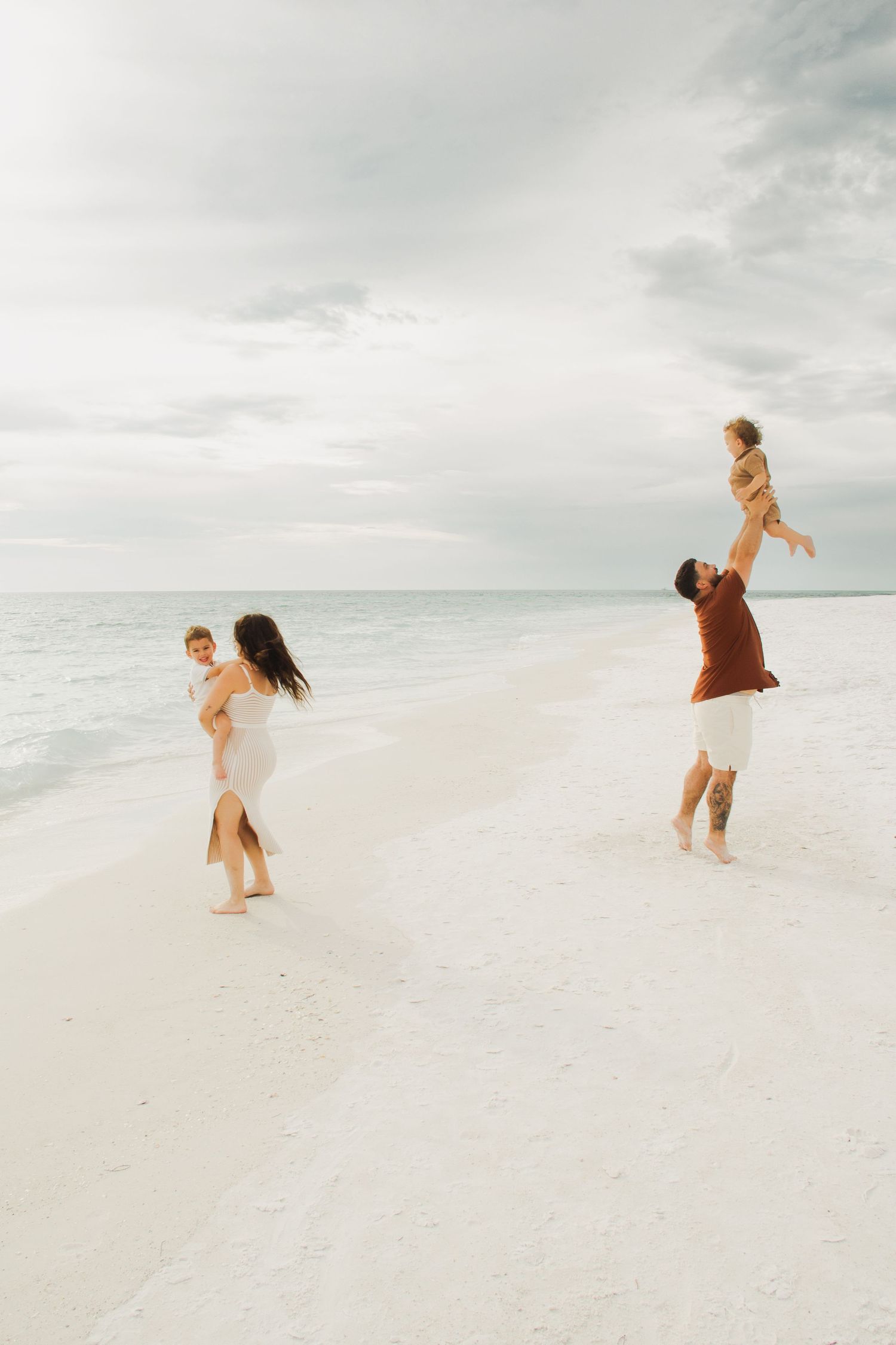 Family playing on a white sand beach, parents lifting children with calm ocean and cloudy sky backdrop.