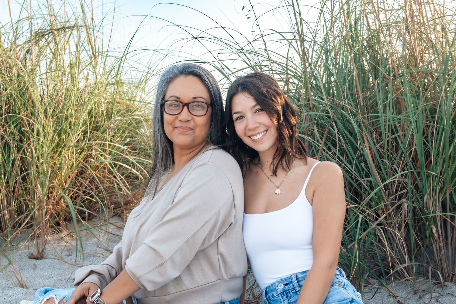 Two women smiling together on a sandy beach surrounded by tall grass on a sunny day.