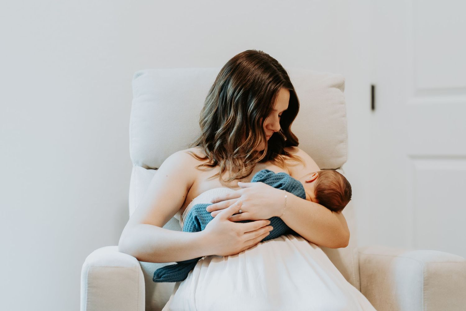 A mother sits in a cozy chair, tenderly cradling and breastfeeding her newborn baby wrapped in a blue blanket.