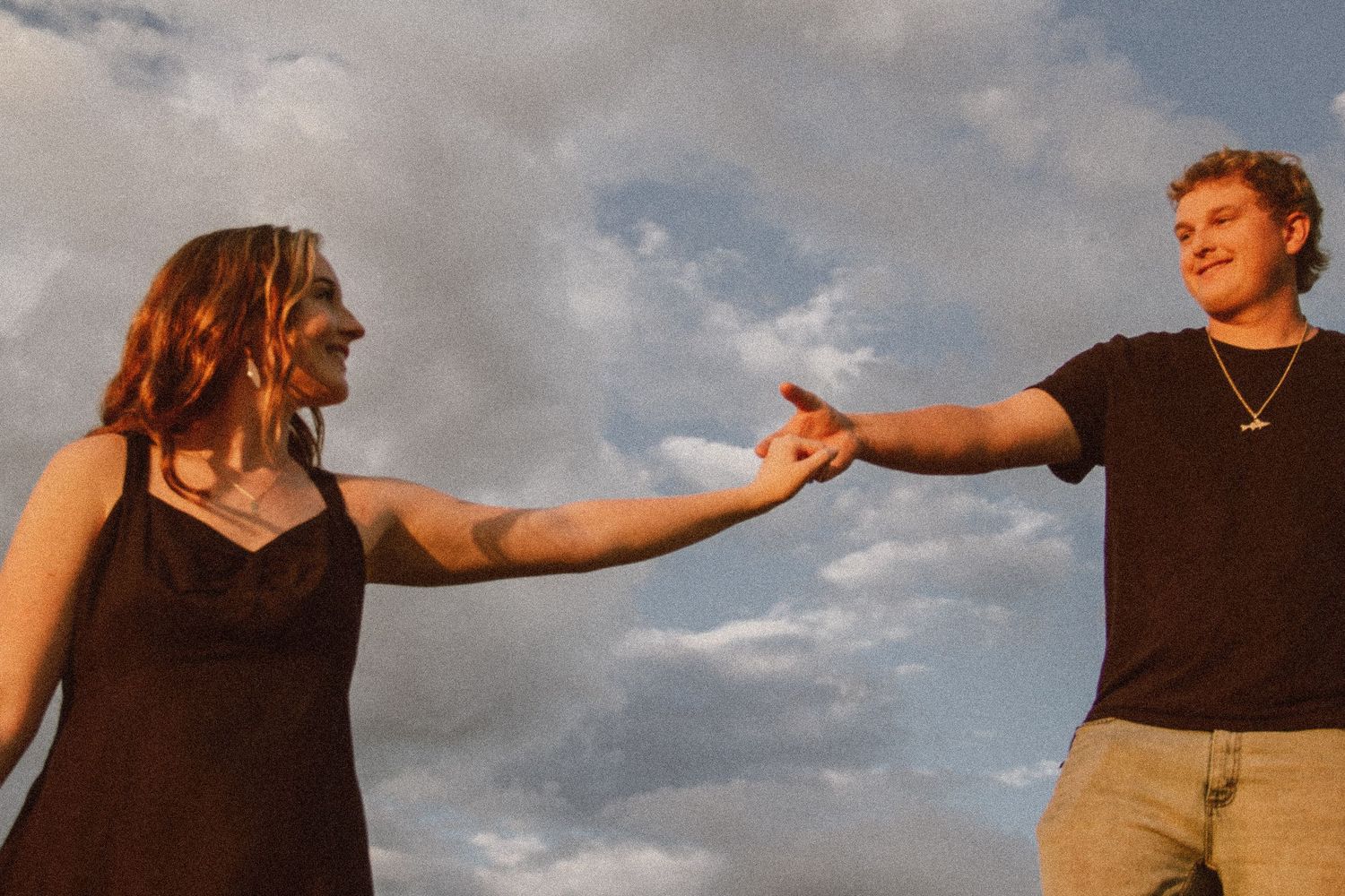 A young couple reaching out to hold hands against a dramatic cloudy sky backdrop outdoors.