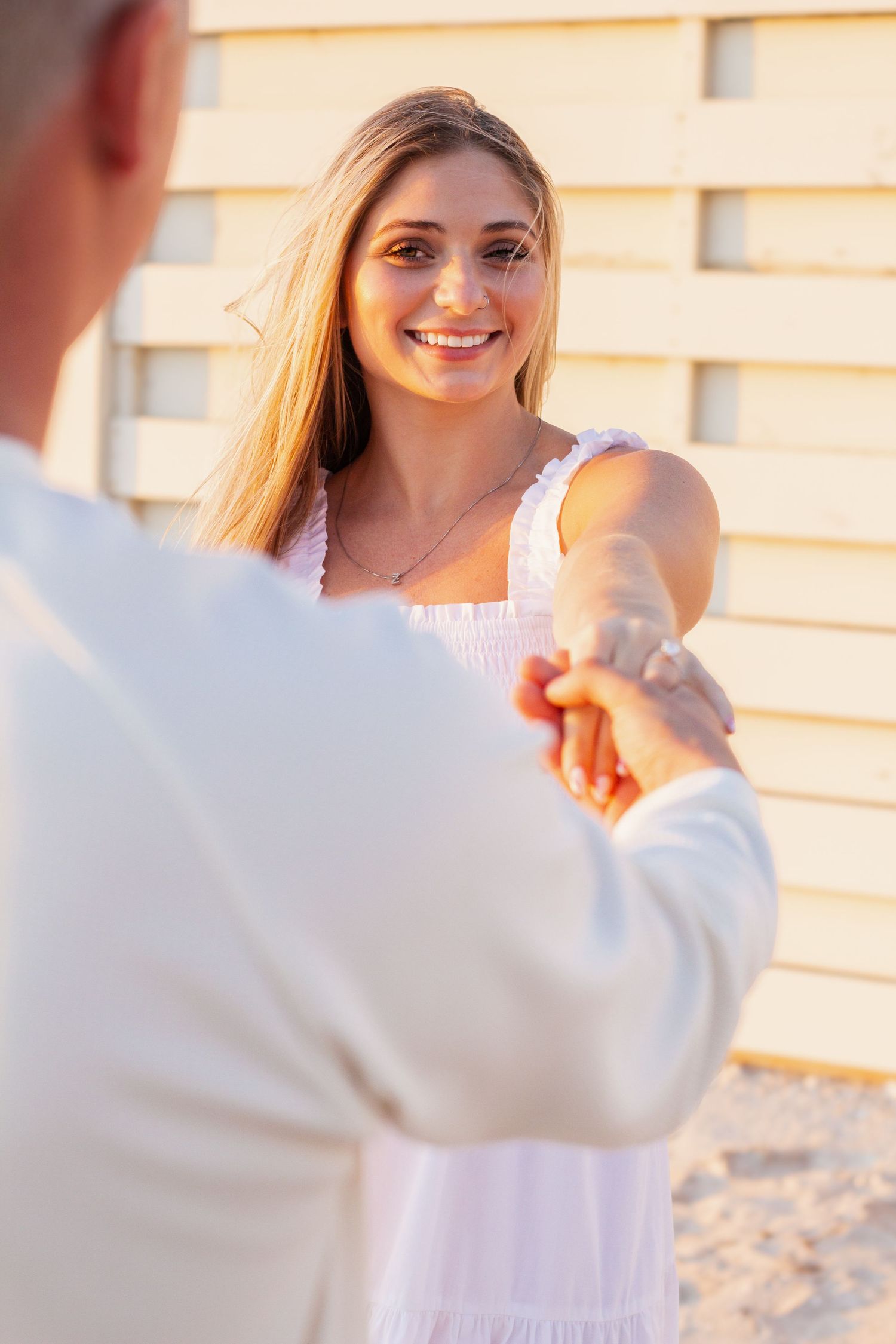 Smiling blonde woman in white dress holds hands with partner outdoors in warm golden sunlight near a wooden wall.