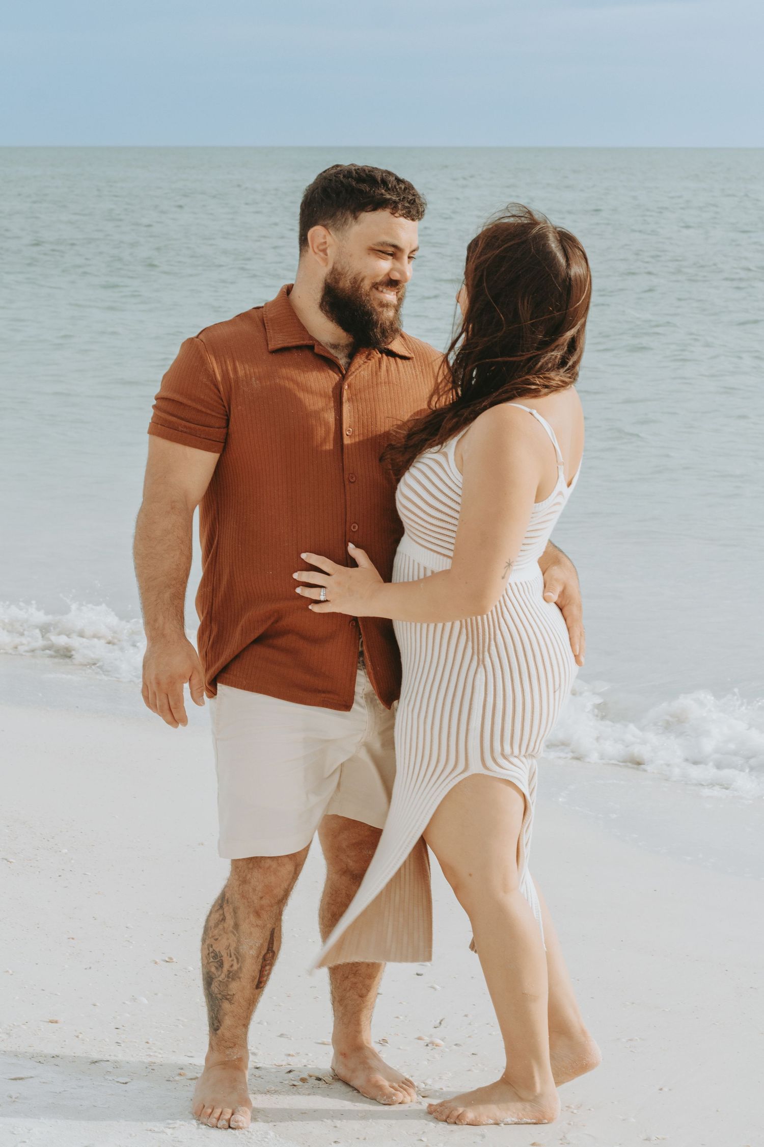 A couple embraces and smiles at each other on a sandy beach, man in rust shirt and woman in striped dress.