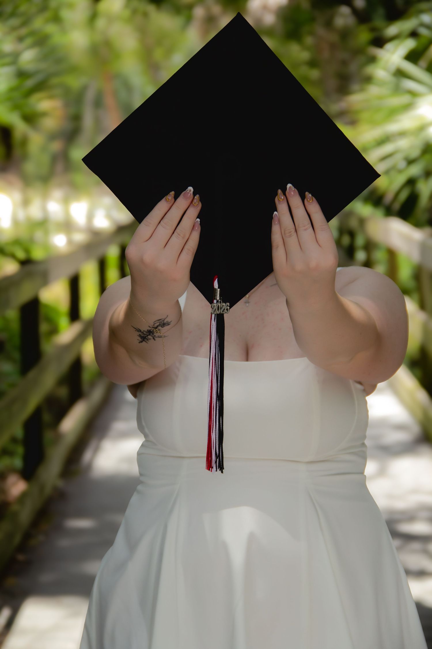 Graduate in white dress holds black mortarboard cap in front of face, showing tassel and wrist tattoo on wooden bridge.
