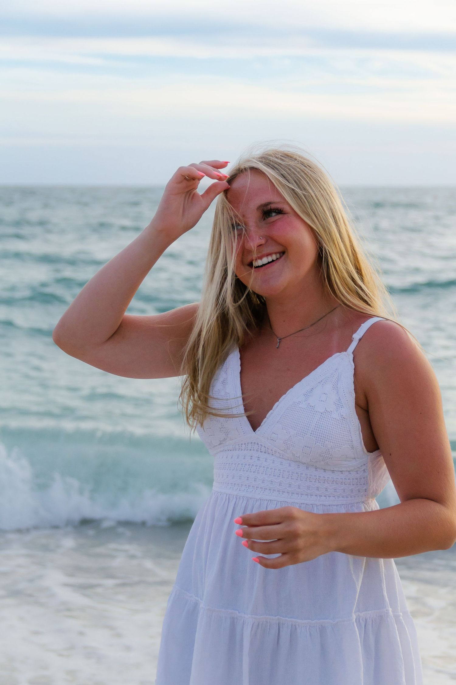 Smiling blonde woman in white sundress posing on a beach with turquoise ocean waves in the background.
