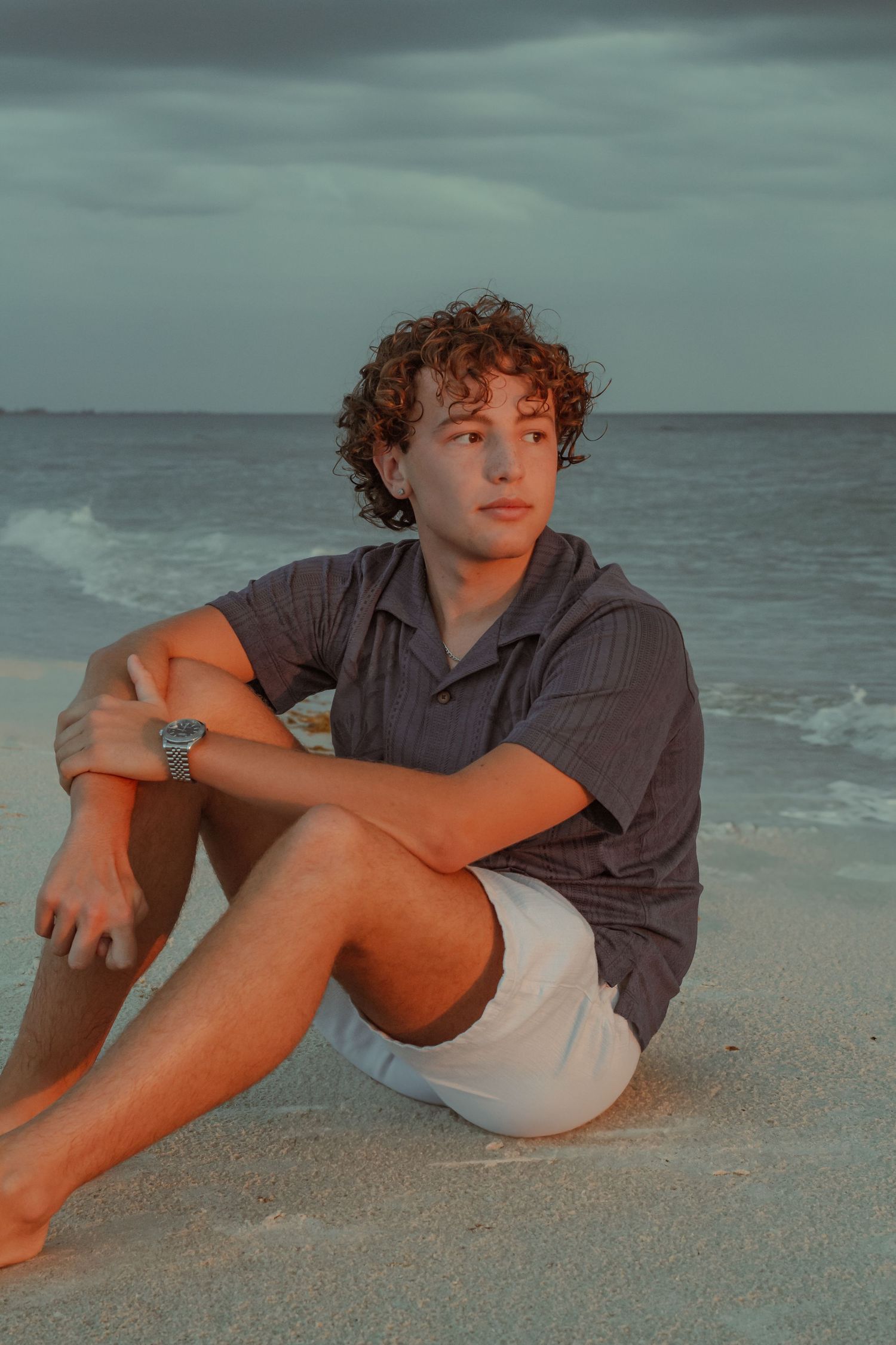 Young curly-haired man in gray shirt and white shorts sitting on sandy beach at golden hour.