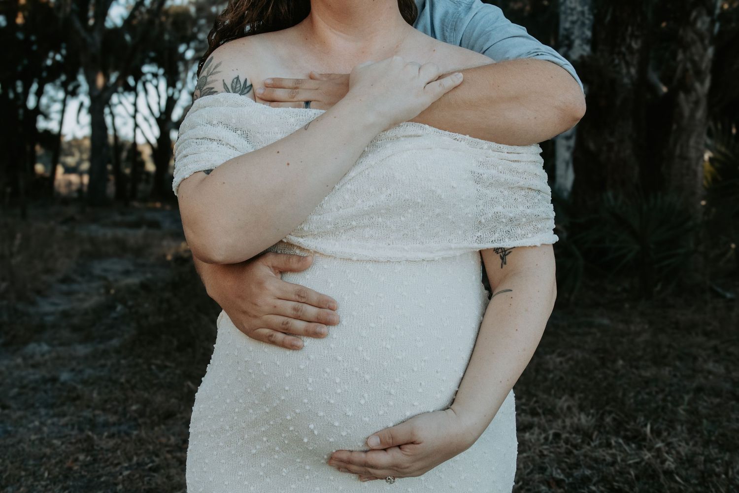 Pregnant woman in white off-shoulder dress being embraced from behind outdoors in a maternity photo shoot.