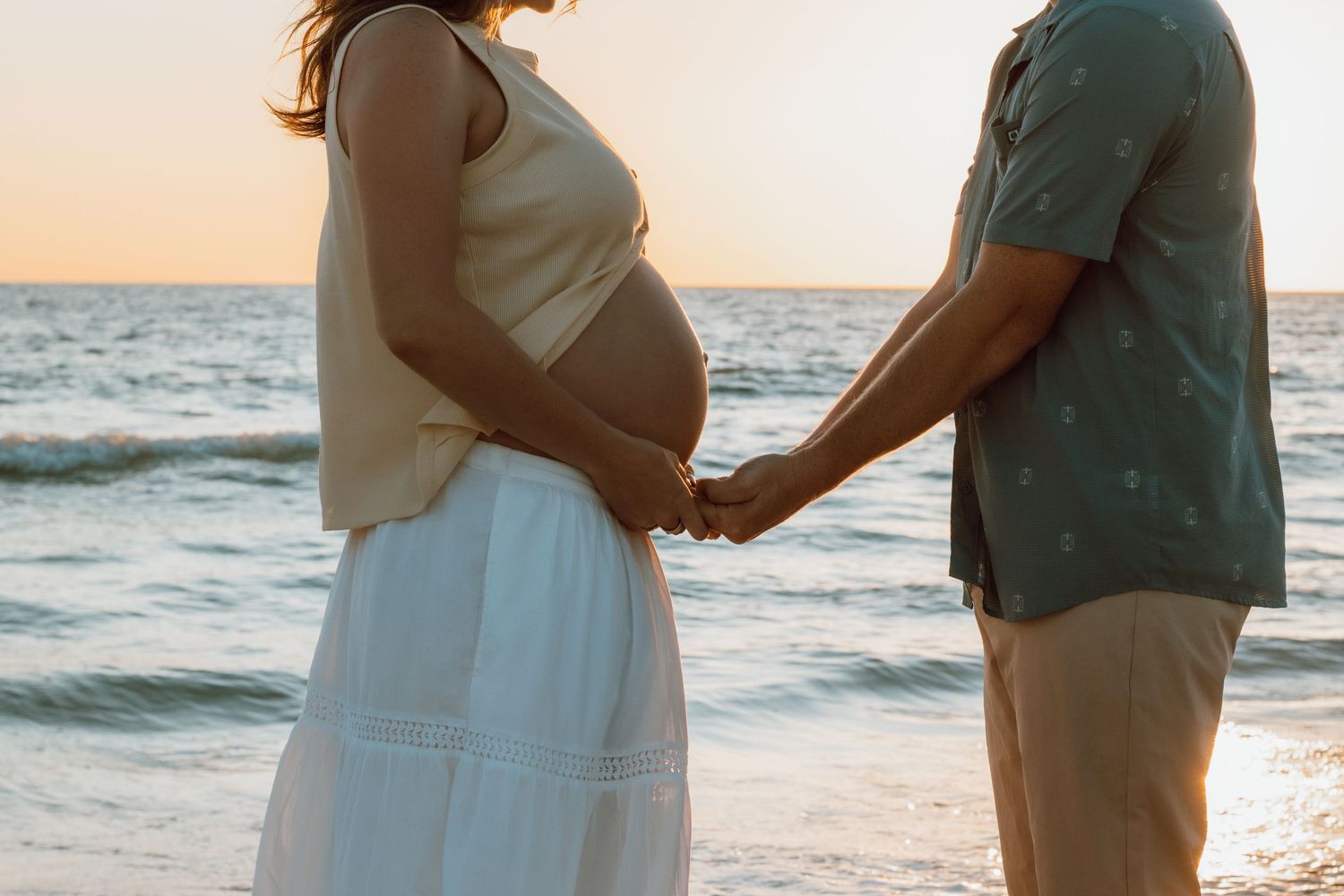 Pregnant couple holding hands on beach at sunset, woman in white skirt showing baby bump by ocean waves.