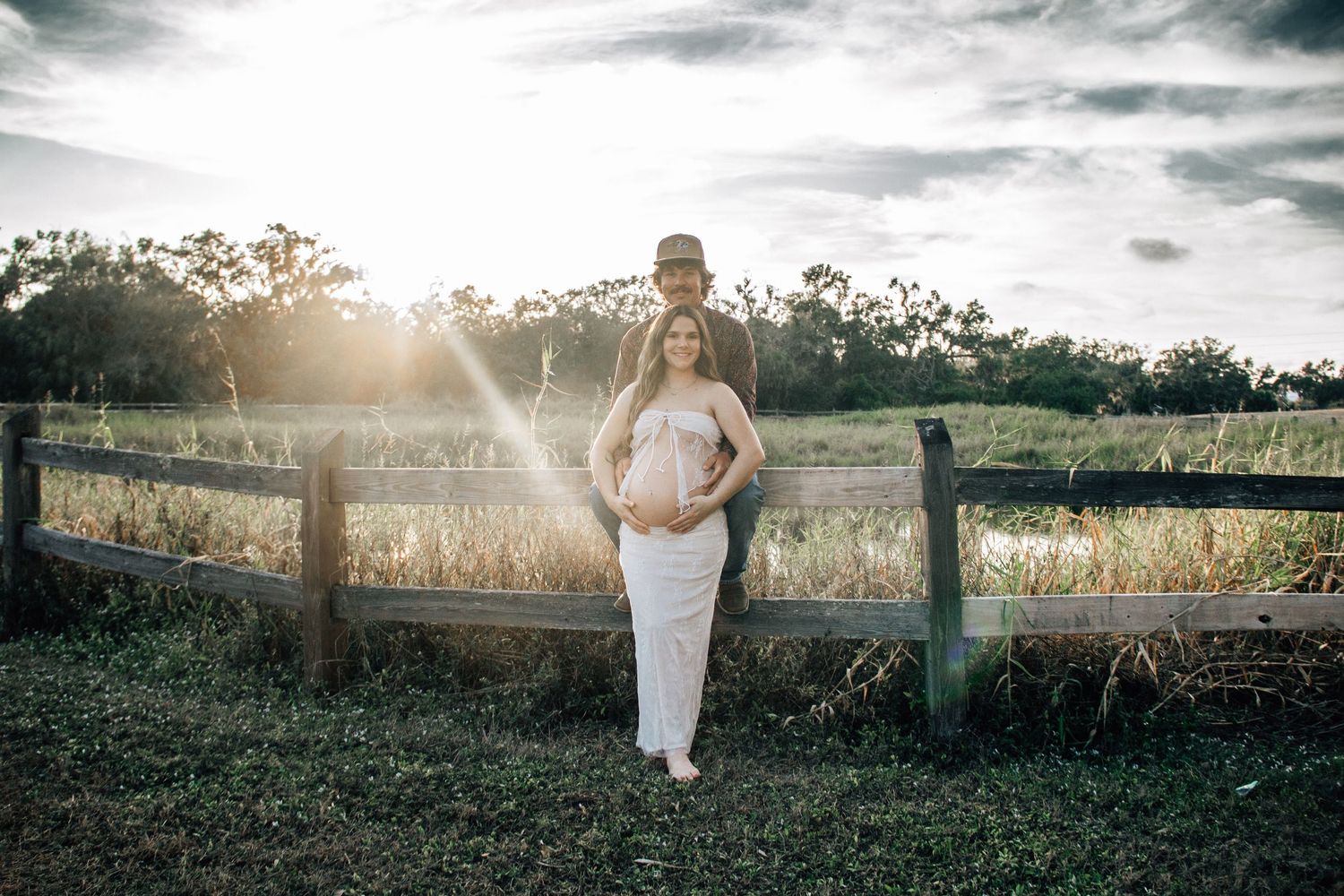Pregnant woman in white dress poses with partner by rustic fence at golden hour in a serene outdoor maternity photo.