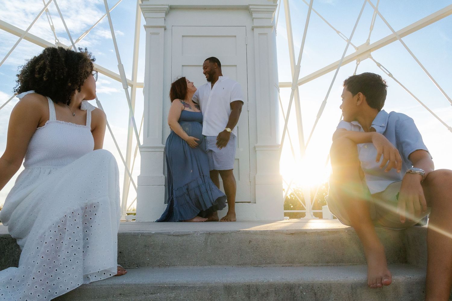 Four people gathered on white steps near a sunlit door, two embracing in the center while others sit and stand nearby.