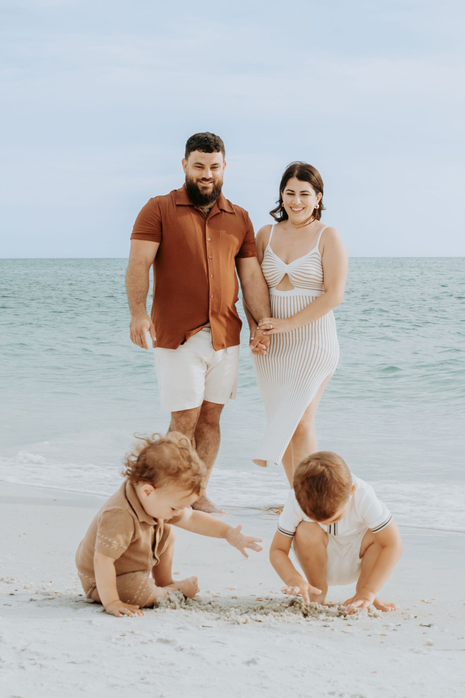 A young family poses on a sandy beach, with two toddlers playing in the sand while parents stand smiling behind them.