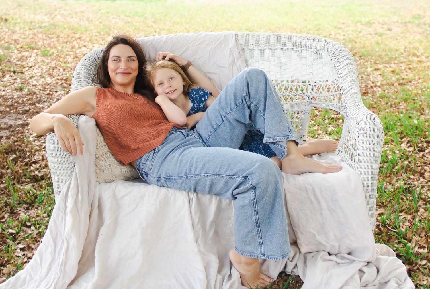 Mother and daughter relaxing together on a white wicker chair outdoors, smiling warmly in casual denim outfits.