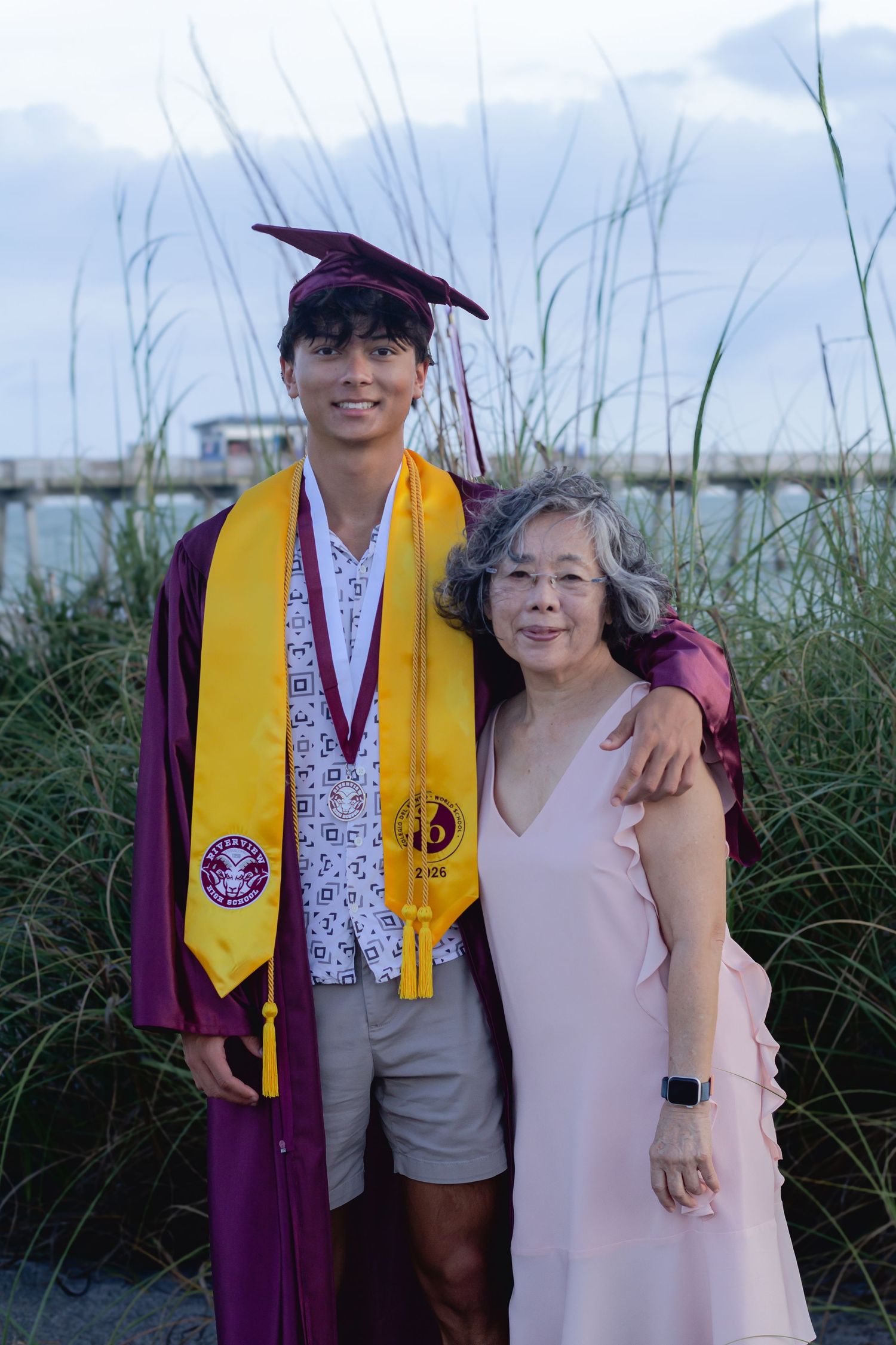 Young man in maroon graduation cap and gown with yellow stole poses with older woman in pink dress outdoors.