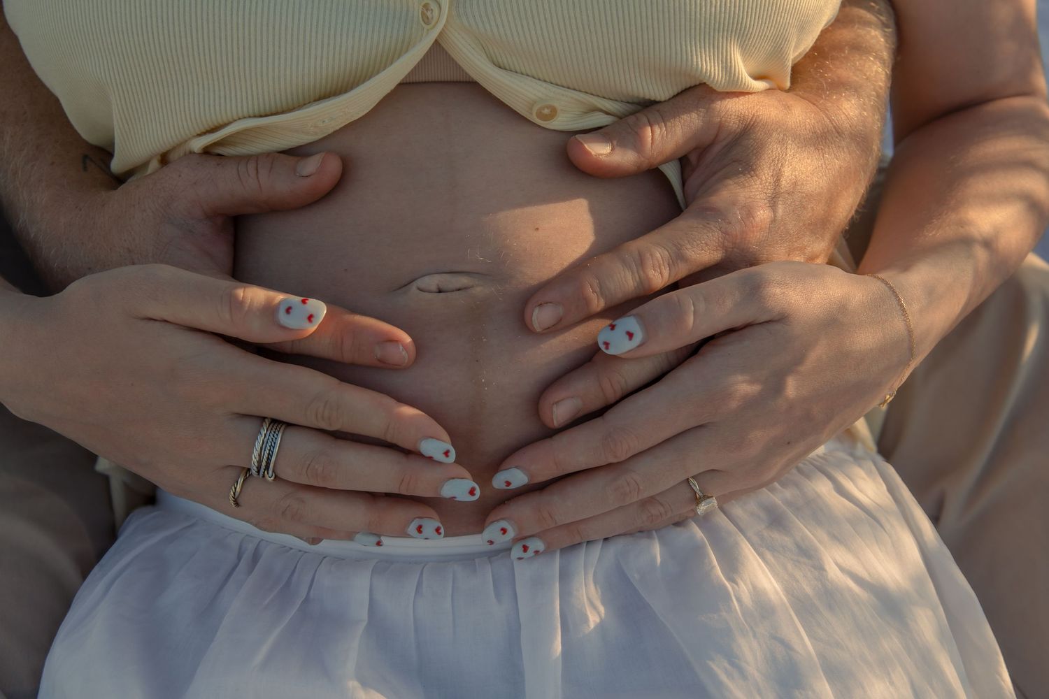 Pregnant woman's bare belly cradled by two pairs of hands, wearing rings, soft warm light.