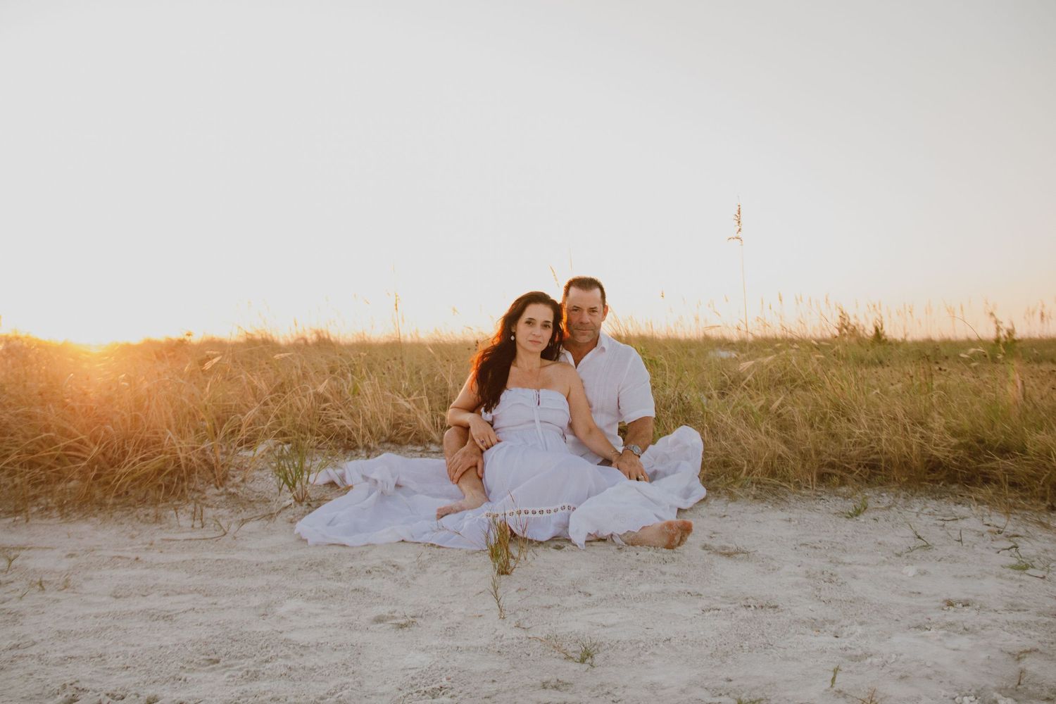 Couple sitting together on sandy beach dunes at golden sunset, woman in white dress, man in white outfit.