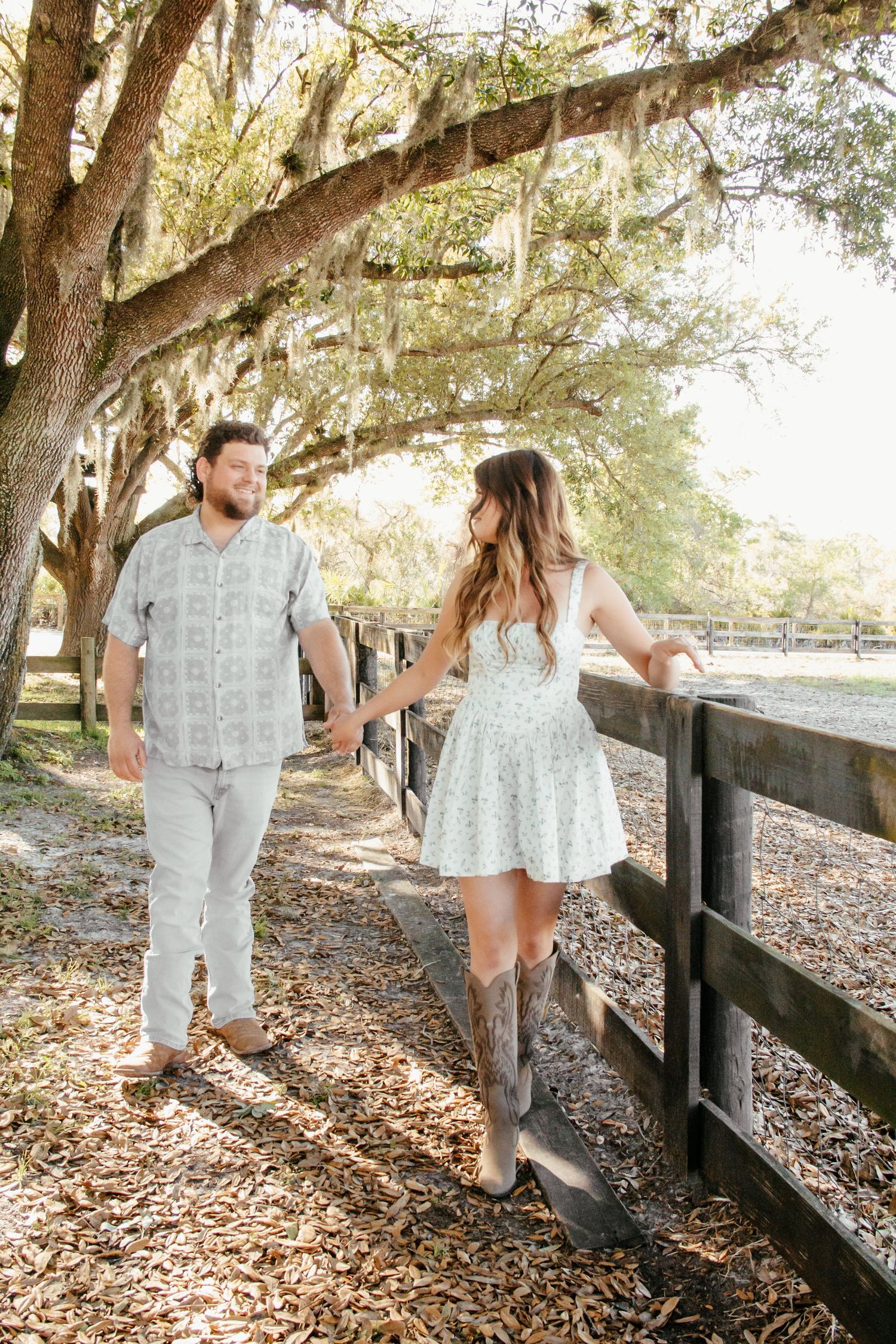 Couple holding hands walking along a wooden fence under moss-draped oak trees on a sunny day.