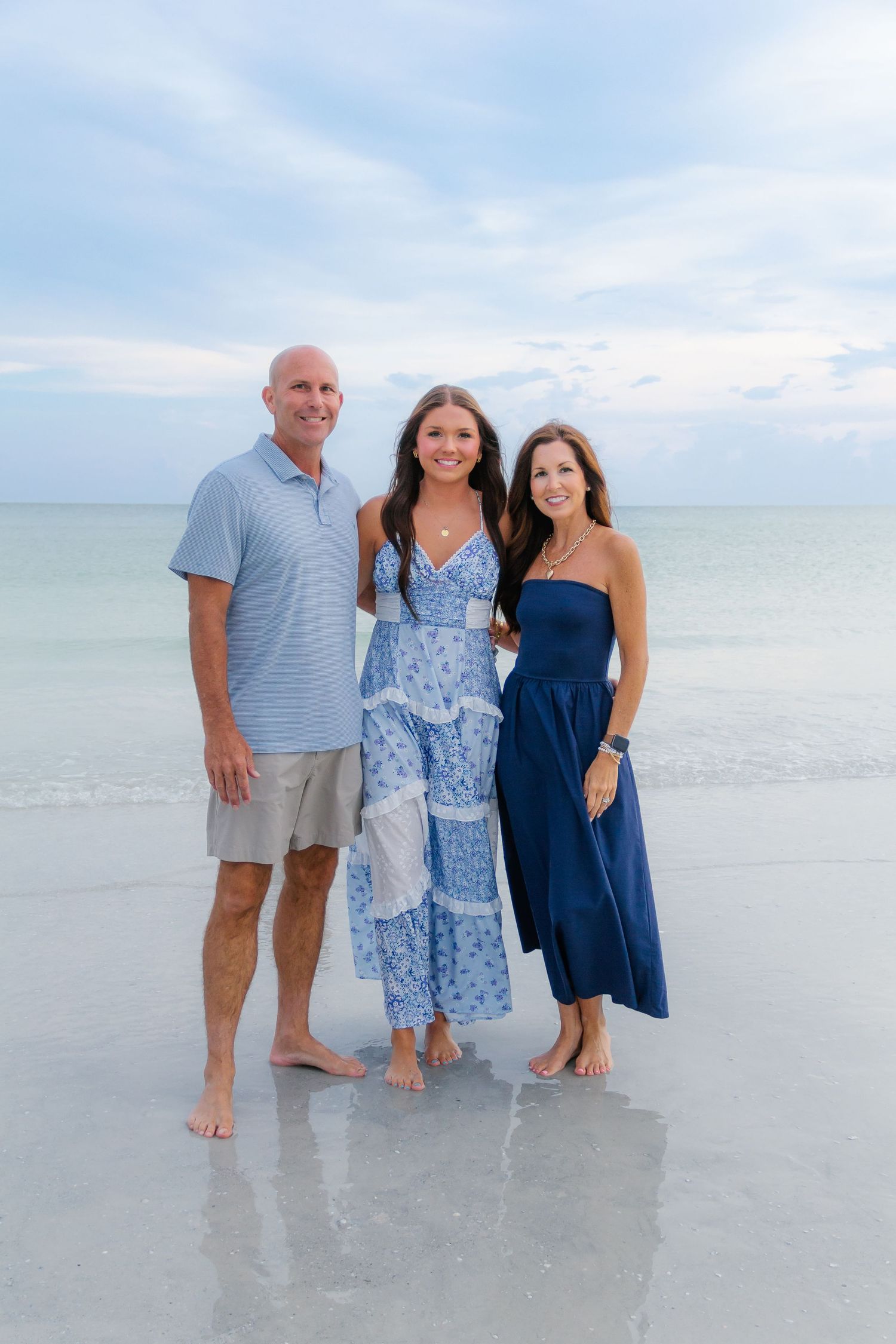Family of three posing barefoot on a serene beach, wearing coordinating blue outfits with calm ocean waters behind them.