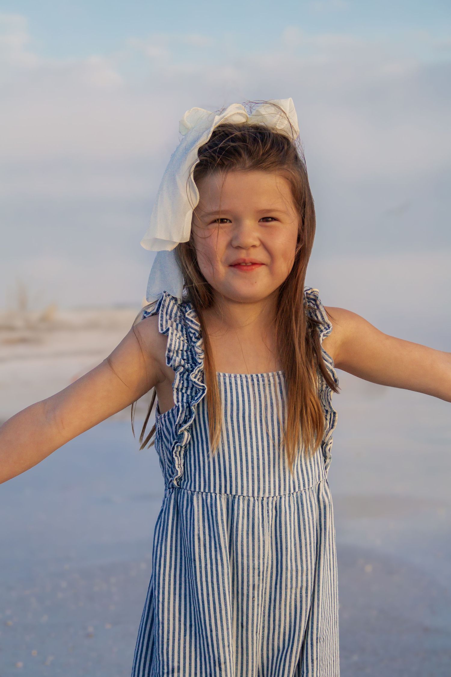 Young girl in blue striped dress with white bow smiles at beach, arms outstretched, golden hour light.