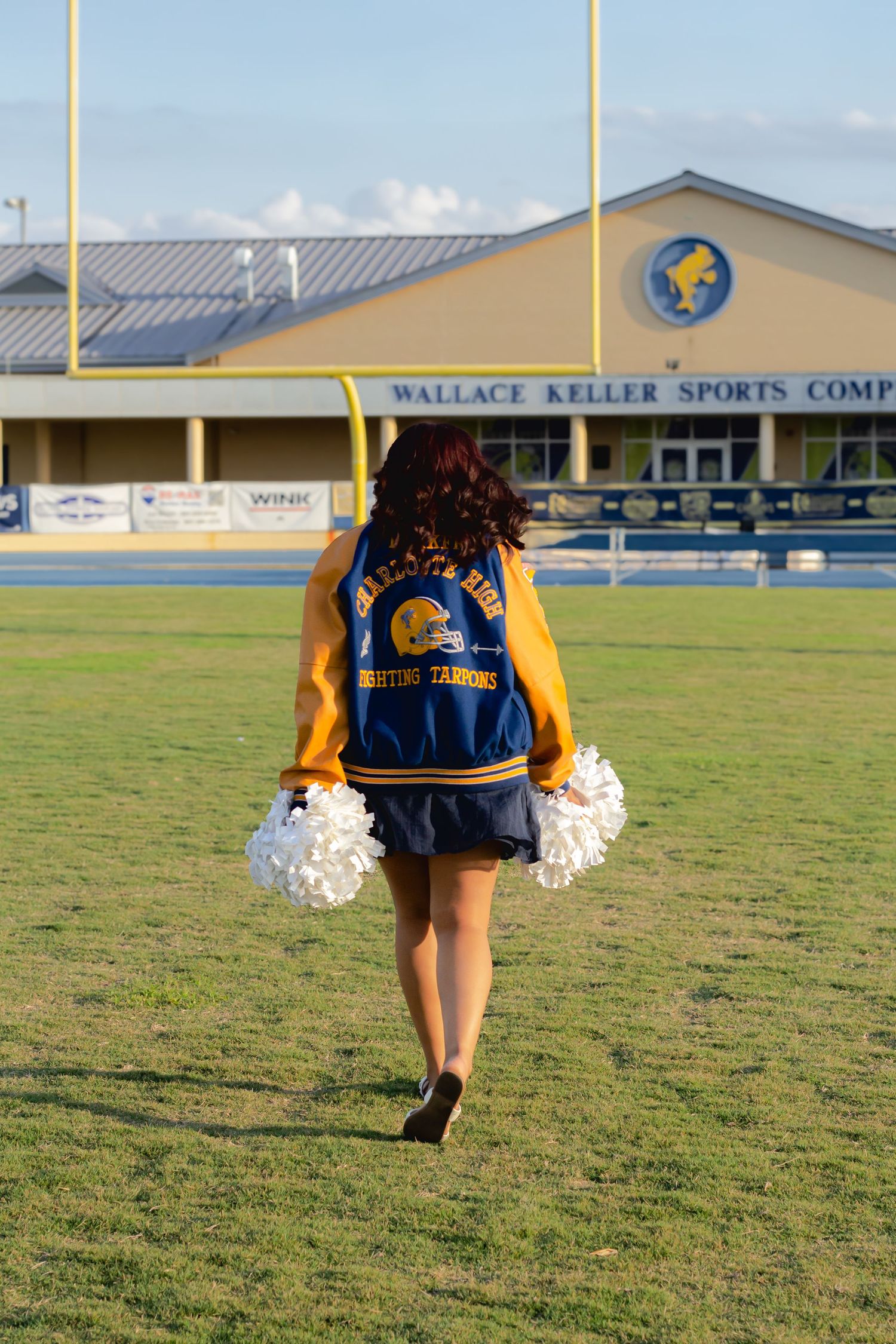Cheerleader in gold jacket with pom-poms walks on football field at Wallace Keller Sports Complex.