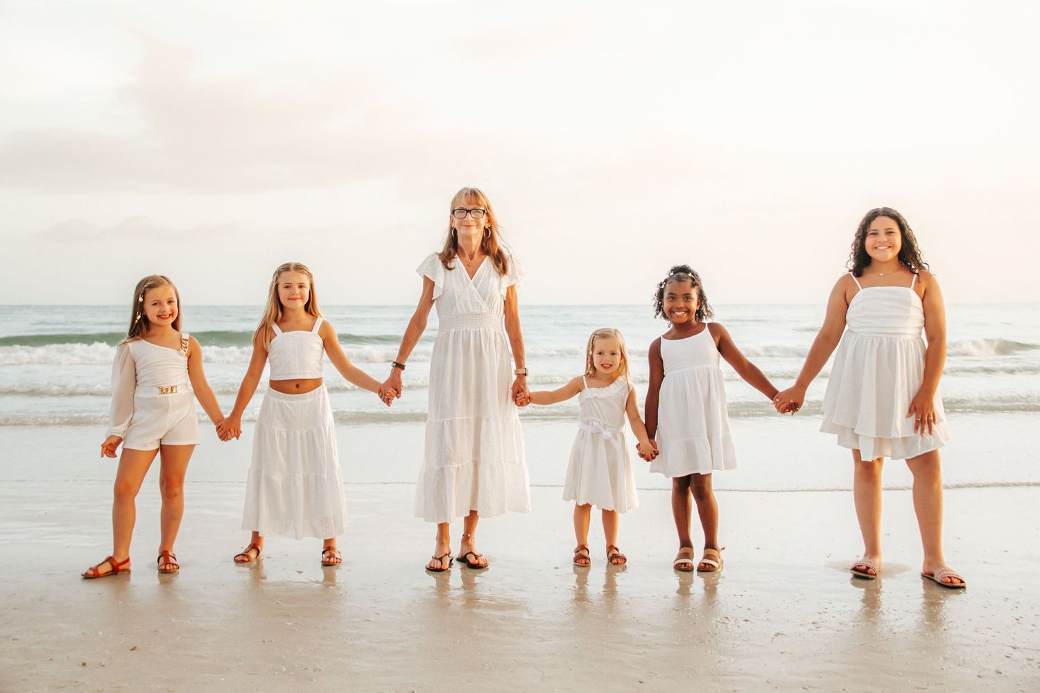 Six girls in white dresses hold hands on a sunny beach with ocean waves in the background.