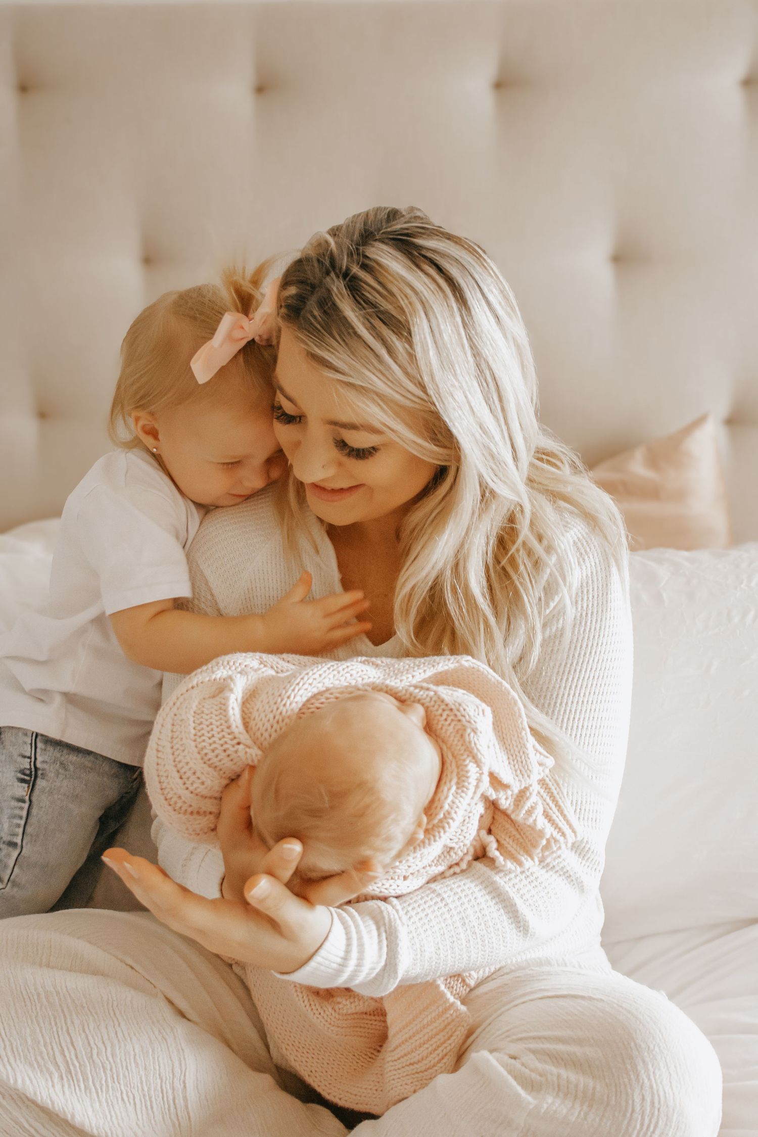 Blonde mother tenderly holding newborn baby while toddler girl with pink bow cuddles her on a white bed.
