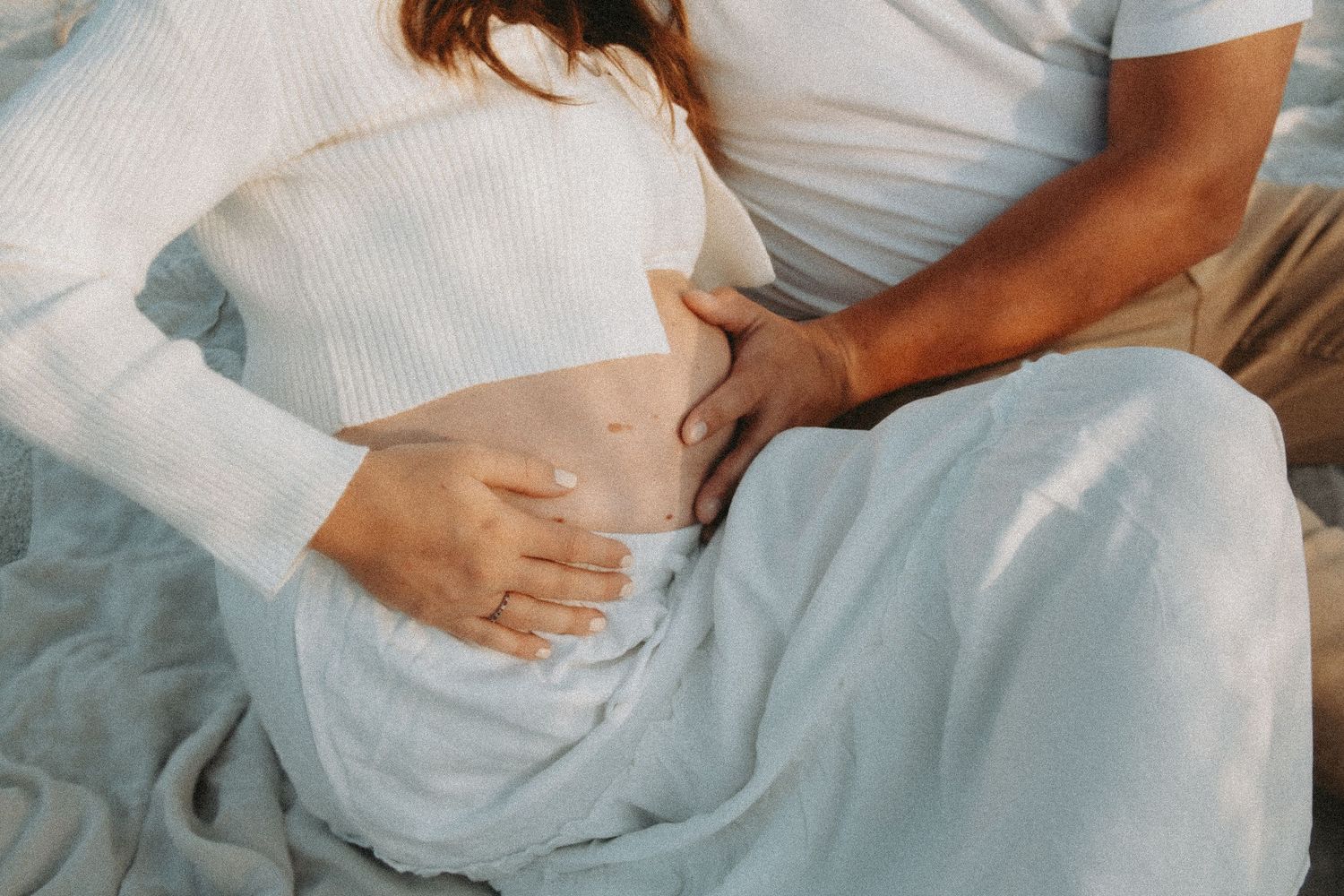 Pregnant woman in white top and blue skirt with partner's hand gently resting on her baby bump.
