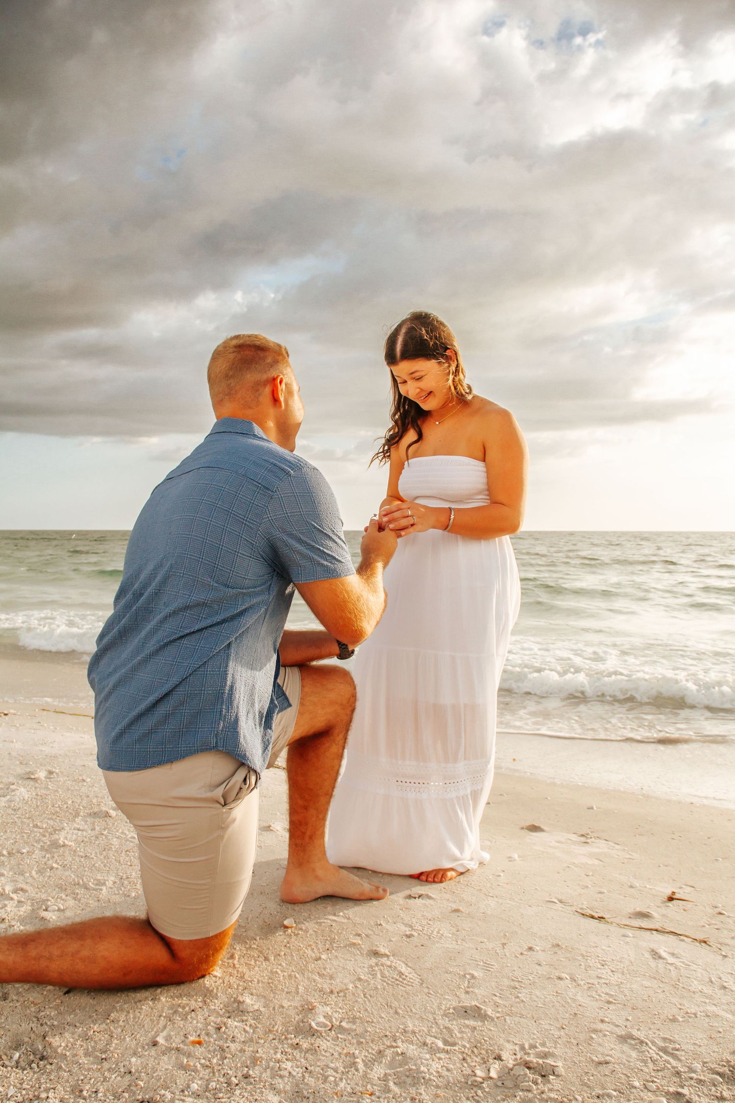Man kneeling on beach proposing to woman in white dress at golden sunset by the ocean.