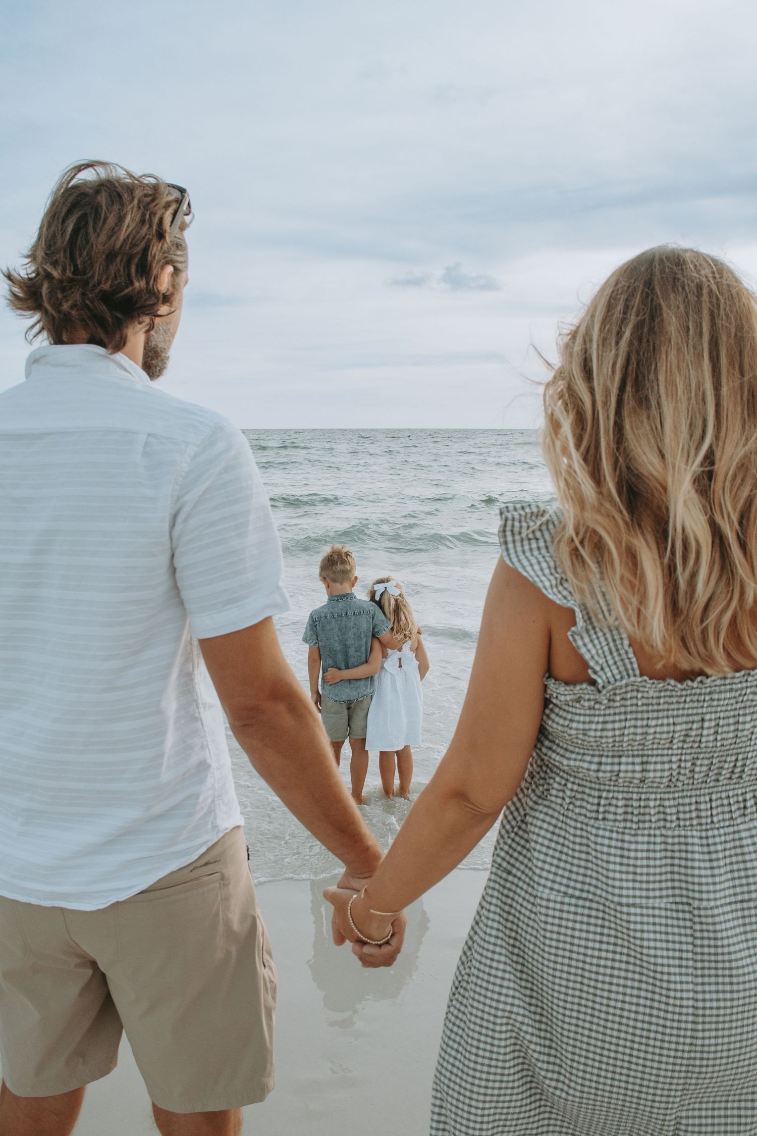 A family holds hands on a sandy beach while two children stand at the ocean's edge on a cloudy day.