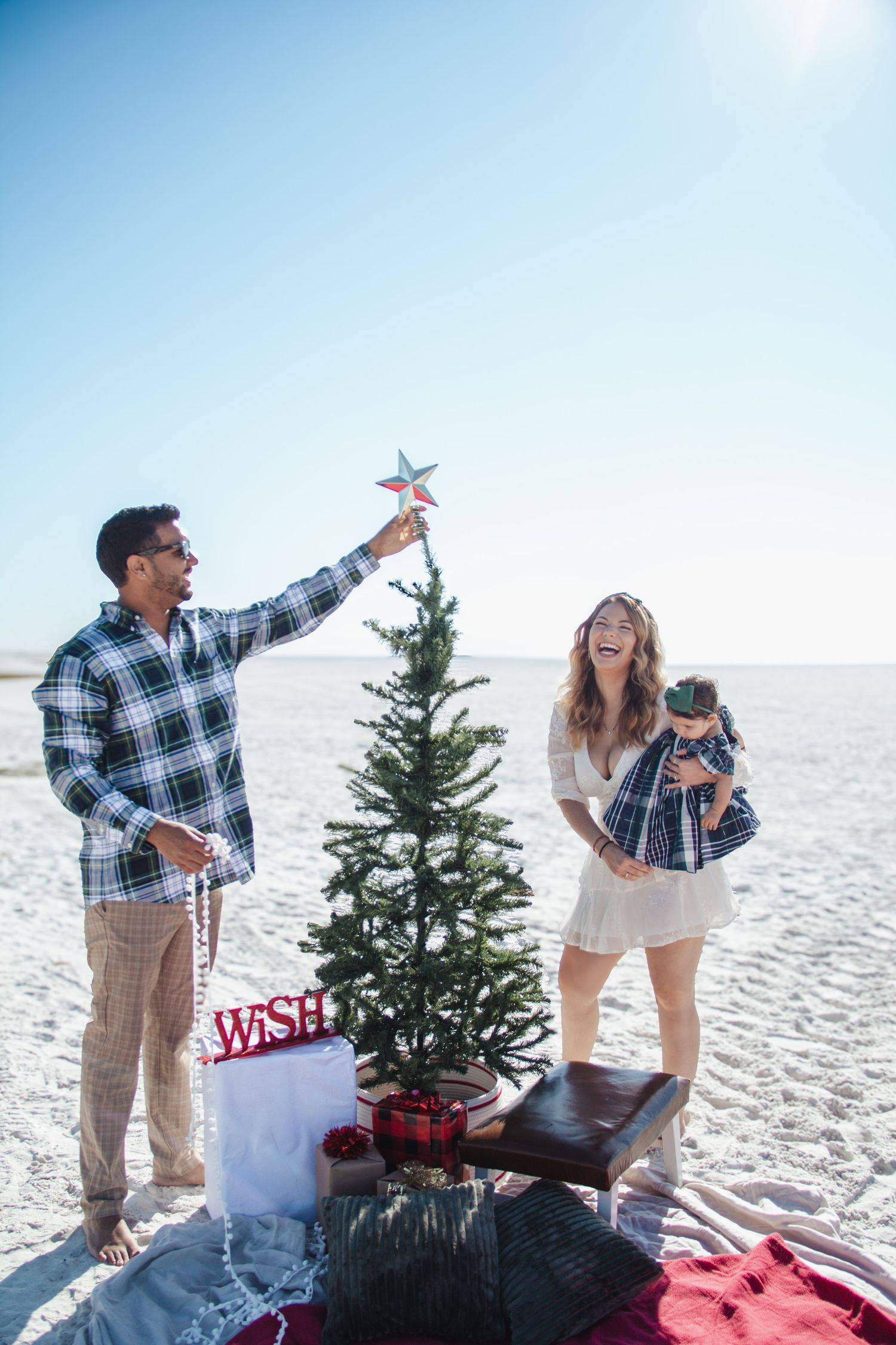 A couple shares joyful moments on a beach with a decorated Christmas tree and holiday decor during a winter photoshoot.