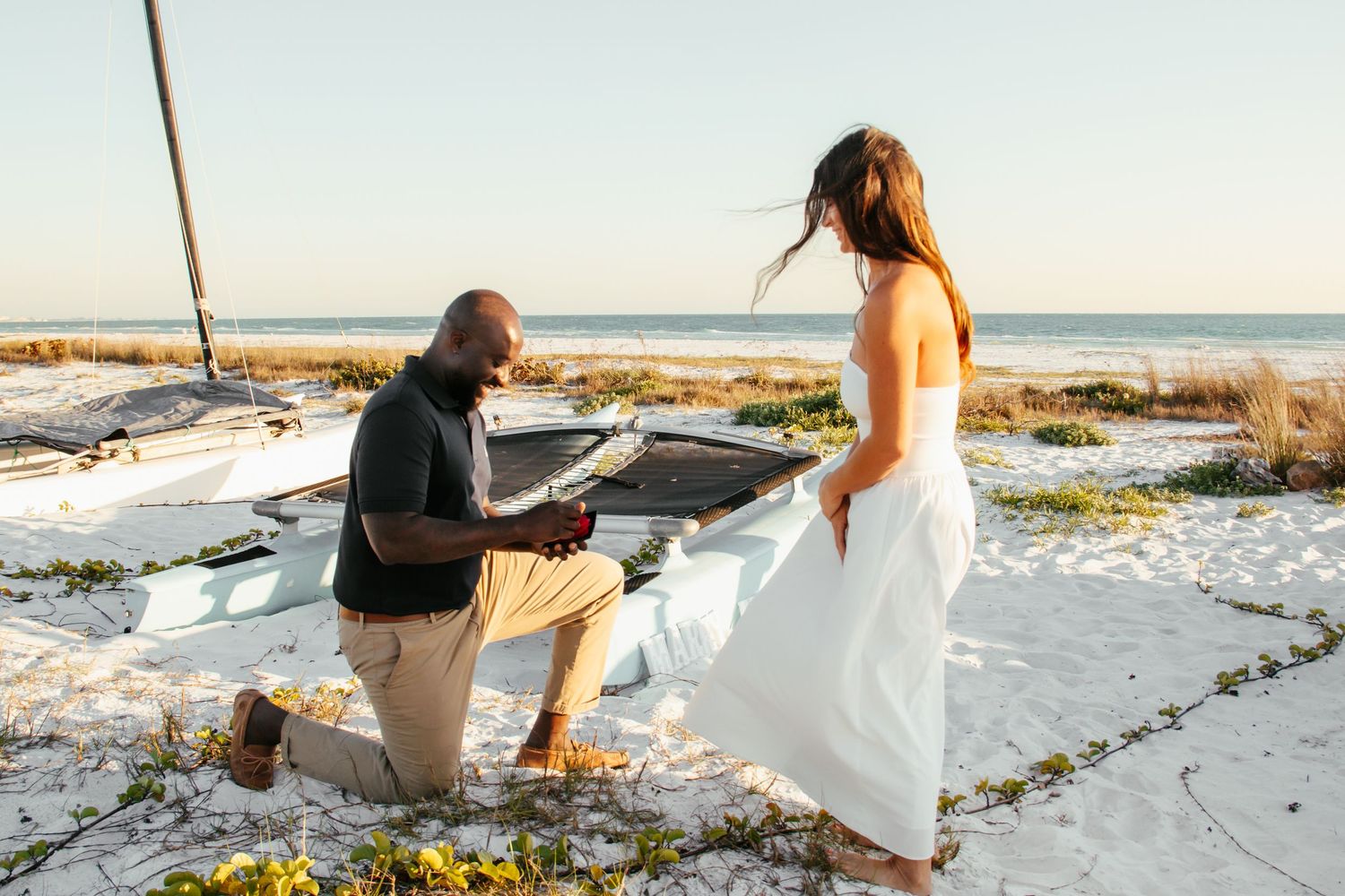 Man kneeling on beach proposing to woman in white dress near ocean at sunset.