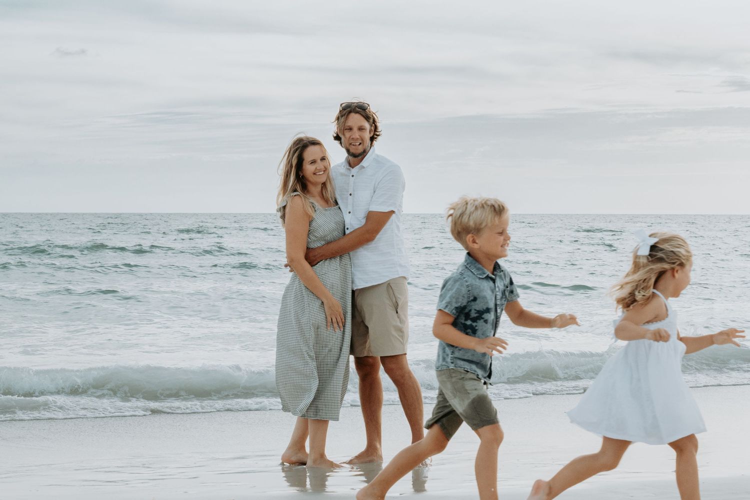 Family of four enjoying a walk on the beach, parents embracing while children run along shoreline.
