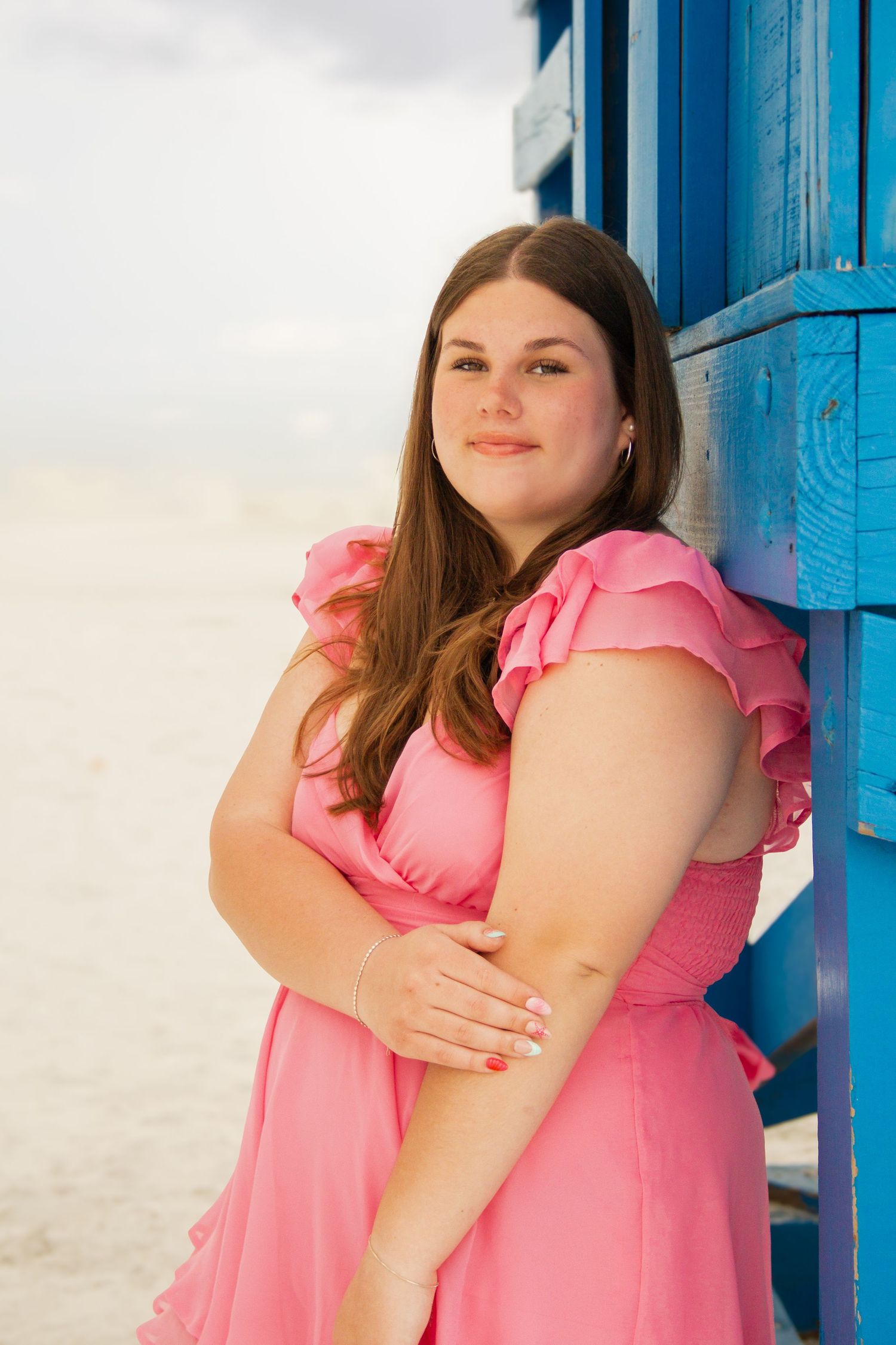 Young woman in pink ruffled dress leaning against blue beach hut, smiling at camera on sandy beach.