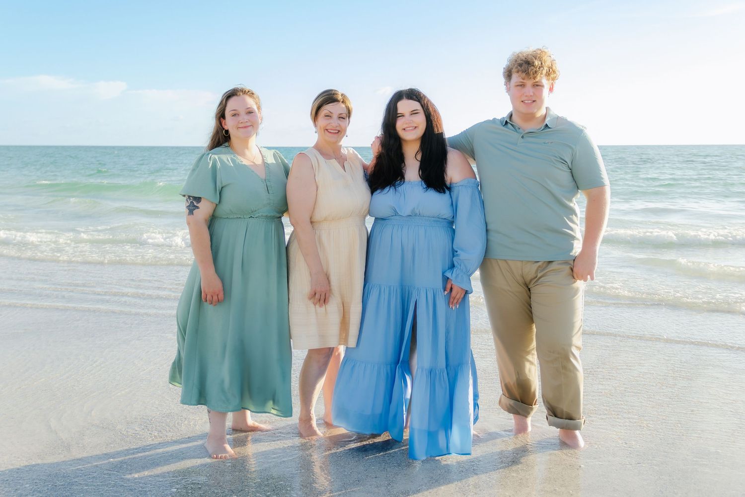 Family of four smiling together on a sunny beach, wearing coordinated outfits in blue and green tones.