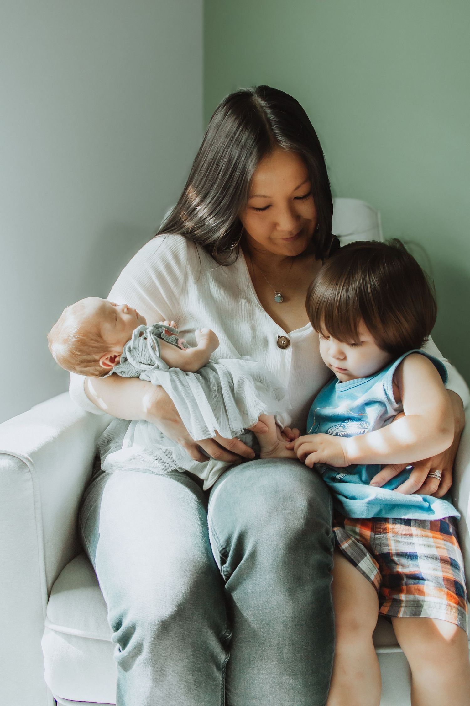 A mother sits in a chair holding a newborn baby while her toddler leans against her in a soft green room.