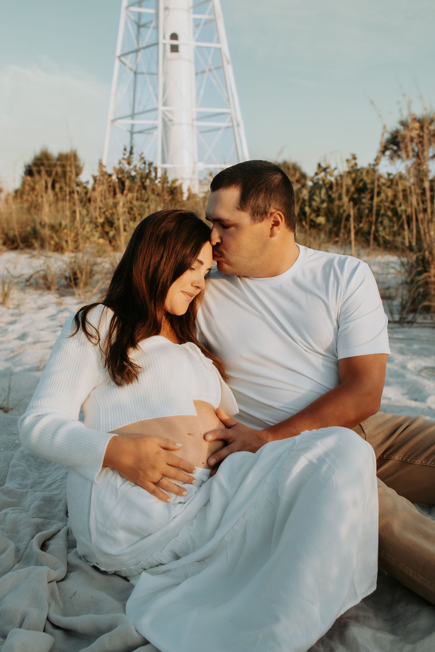 A couple shares a tender moment on a sandy beach, the pregnant woman cradled by her partner near a white lighthouse.